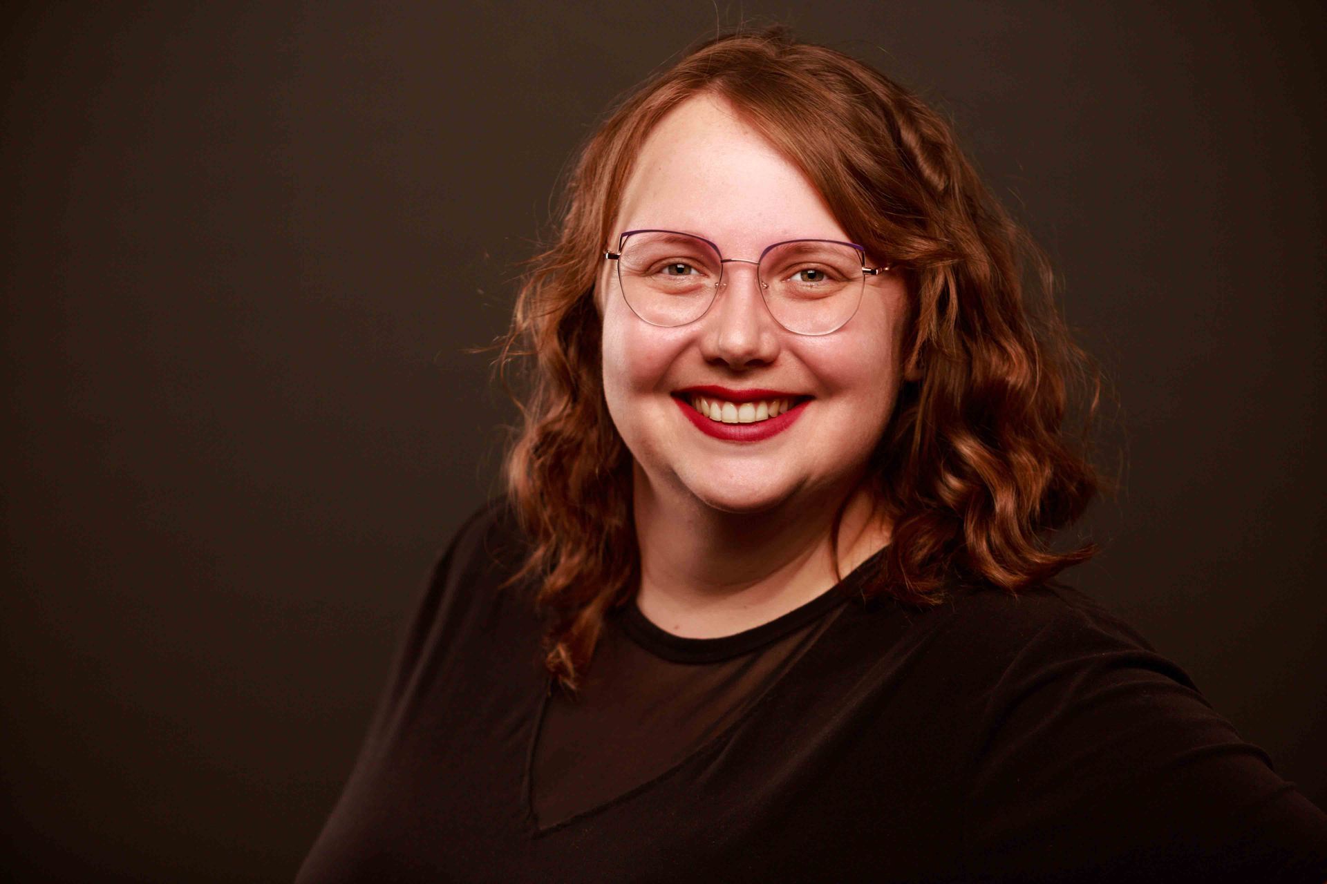 Smiling woman with glasses and wavy brown hair against a dark brown background