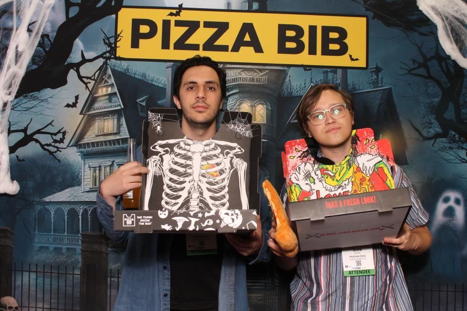 Two people holding pizza boxes under a “PIZZA BIB” sign, posing in a spooky backdrop.