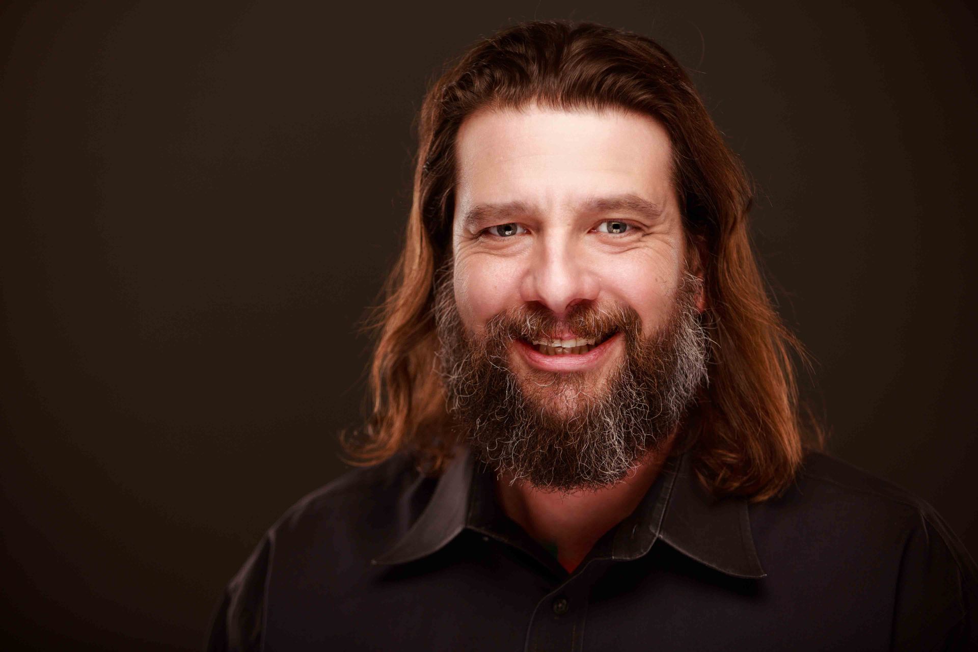 Smiling person with long hair and beard against a dark brown background
