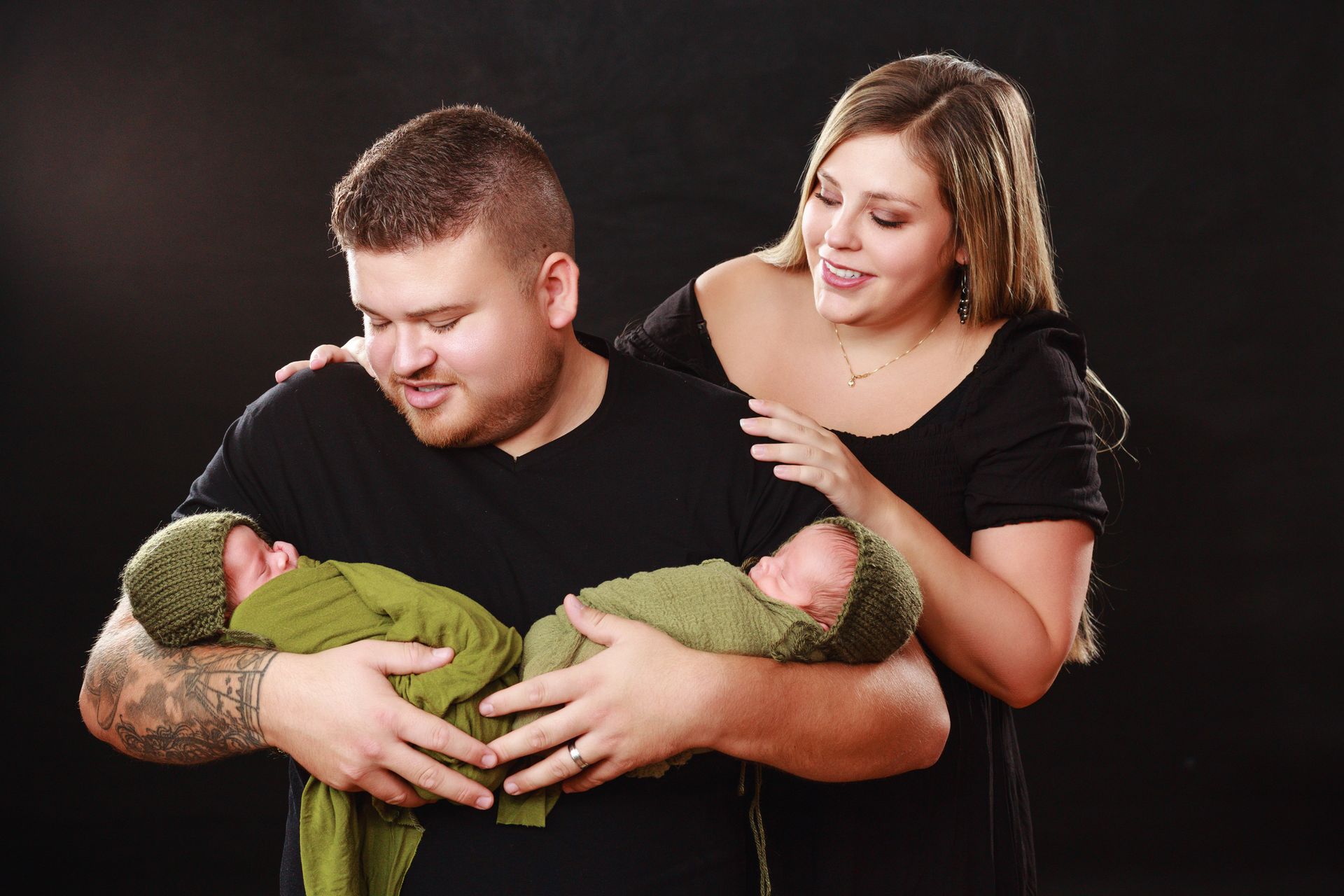 Two adults in black shirts holding two newborn babies wrapped in green blankets against a dark background
