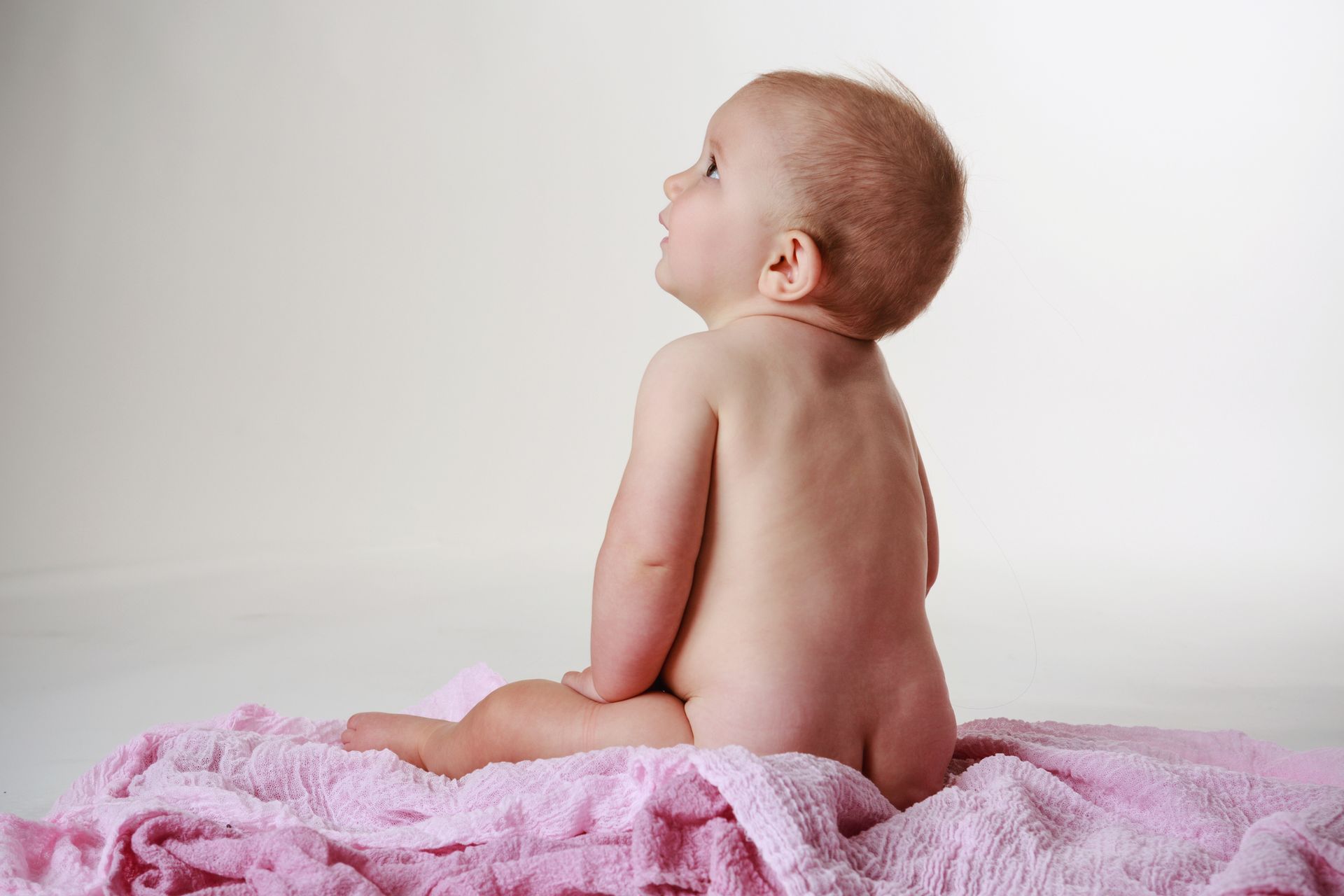 Baby sitting on a pink blanket, looking upward against a plain light background