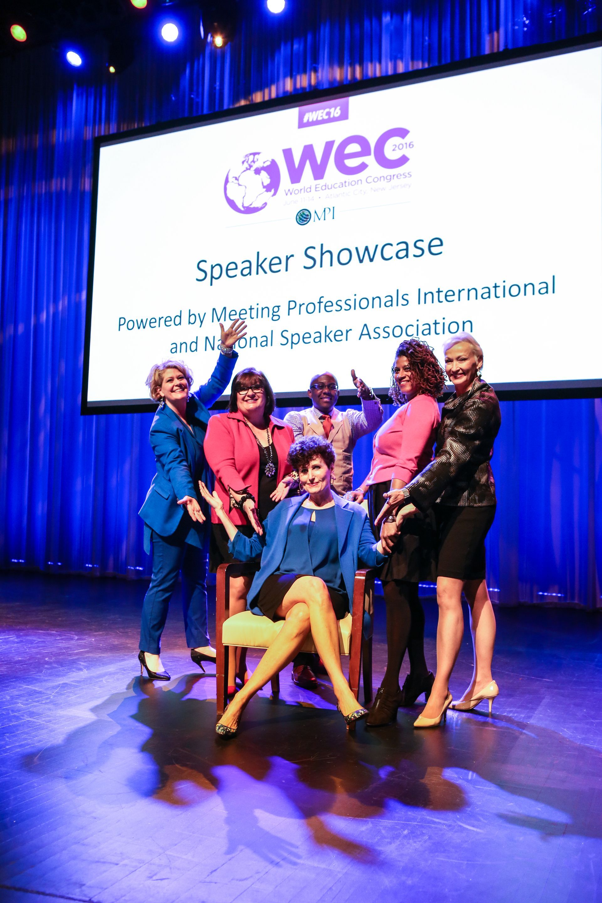 Group of people posing on stage at a WEC Speaker Showcase, with blue curtains and a large presentation screen.