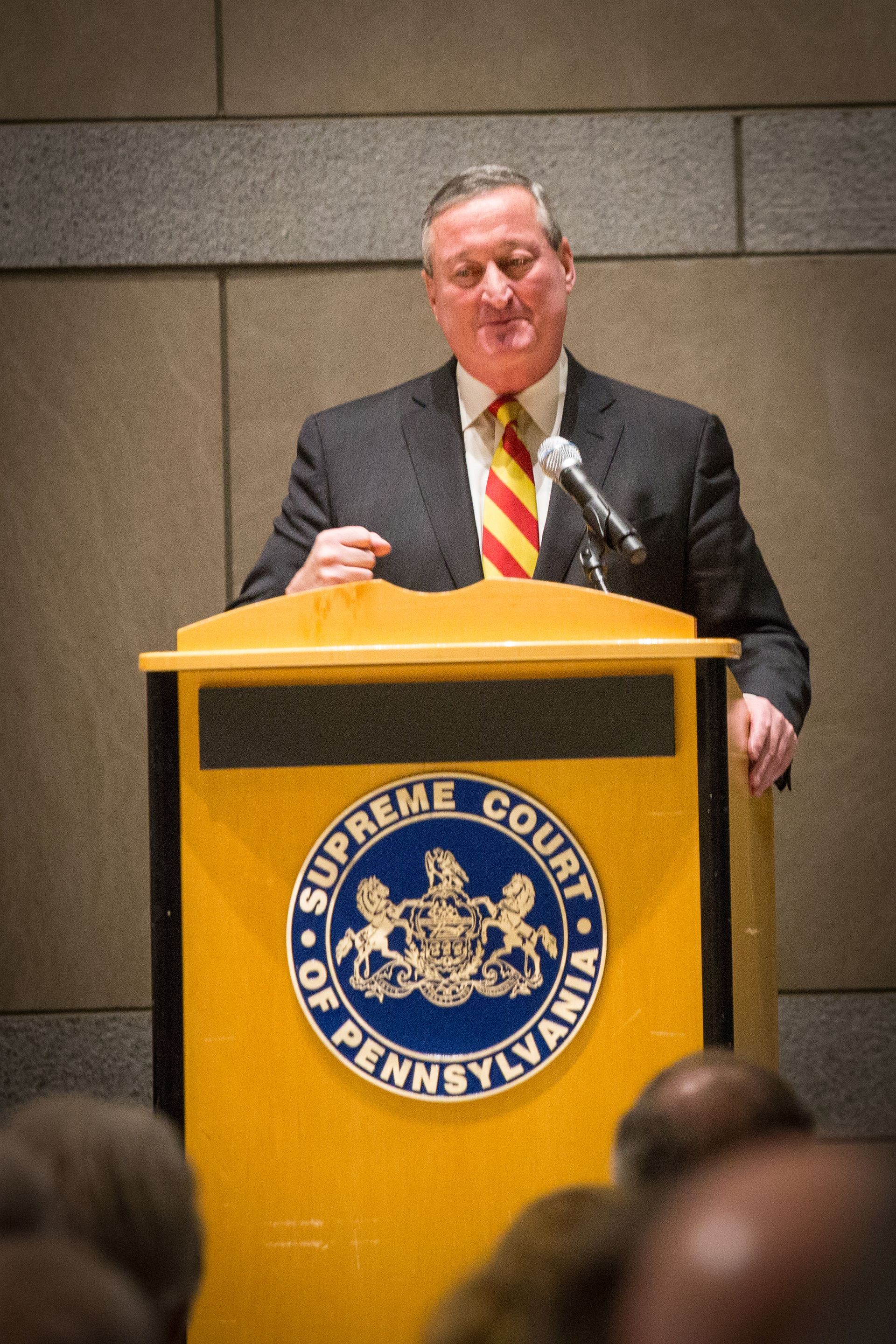 Man speaking at a podium with a blue city seal, wearing a dark suit and red-yellow striped tie