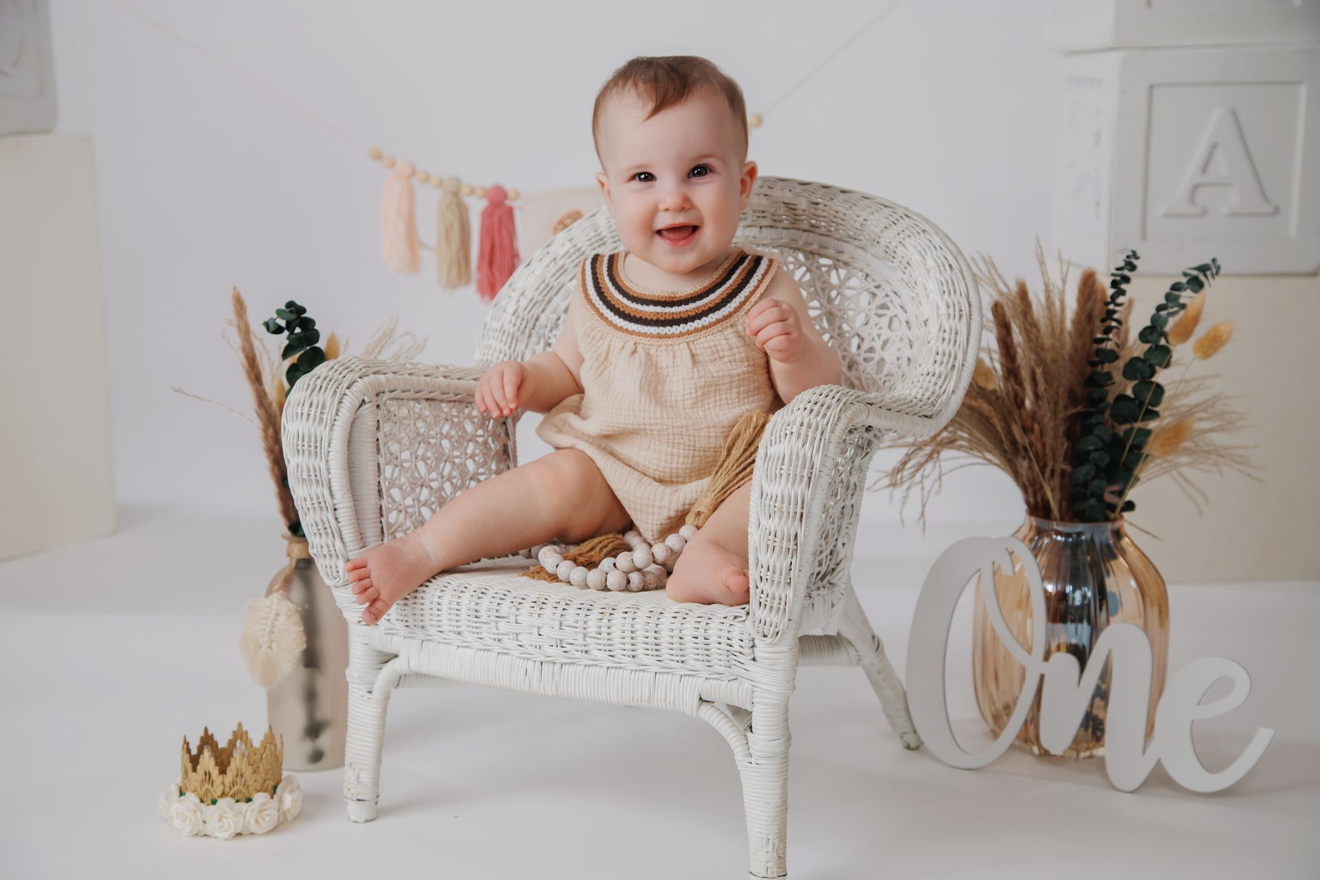 Smiling baby in a cream dress sitting on a white wicker chair with pastel party decor