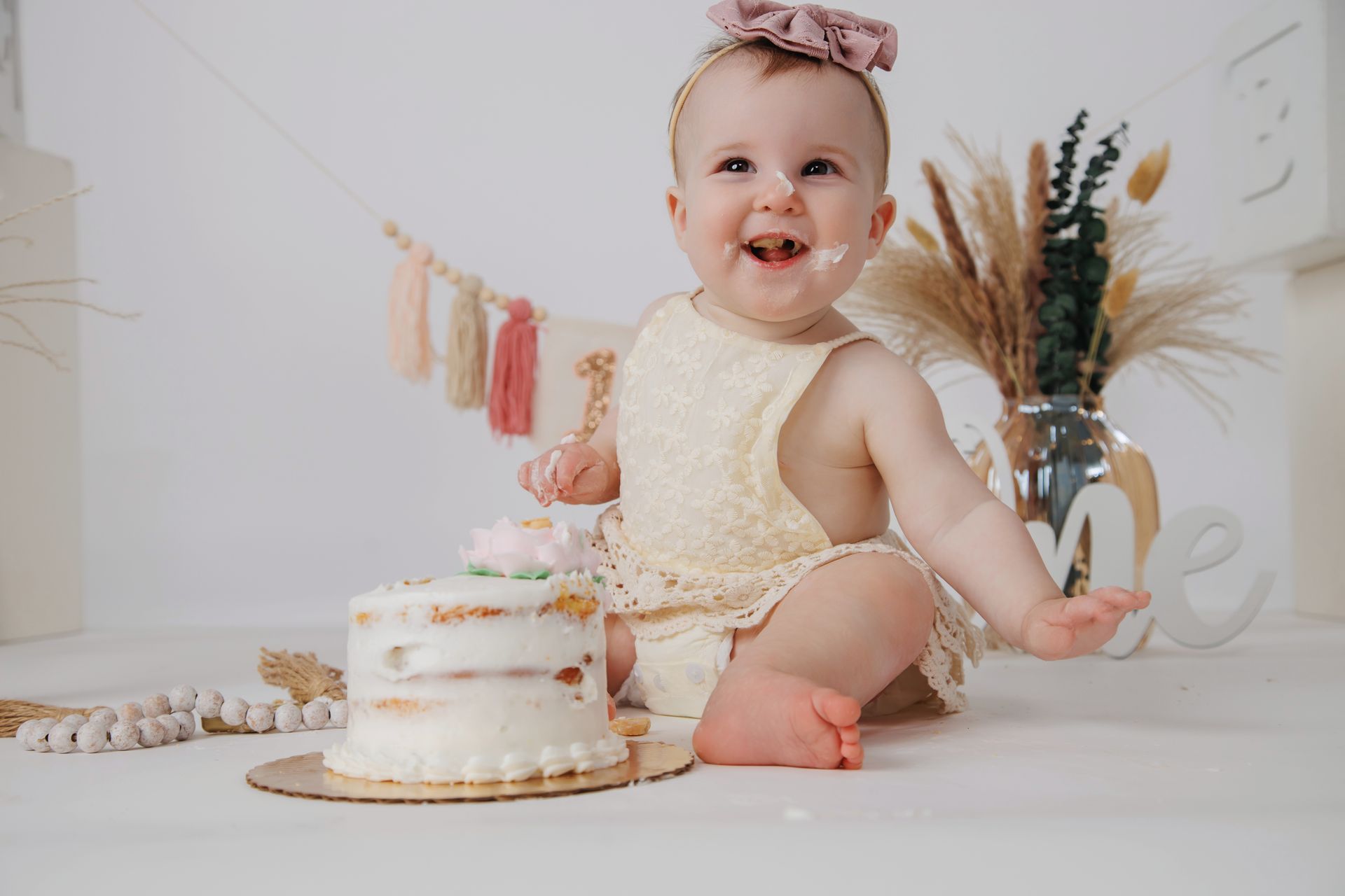 Baby sitting beside a small cake at a pastel first-birthday celebration, smiling in a cream outfit.