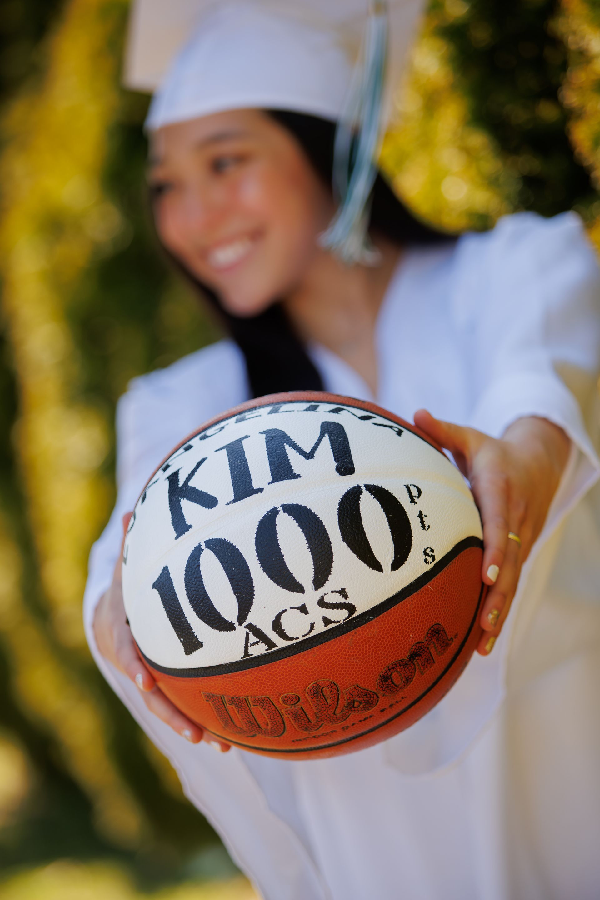 Smiling graduate in white cap and gown holding a basketball with “KIM 1000” written on it