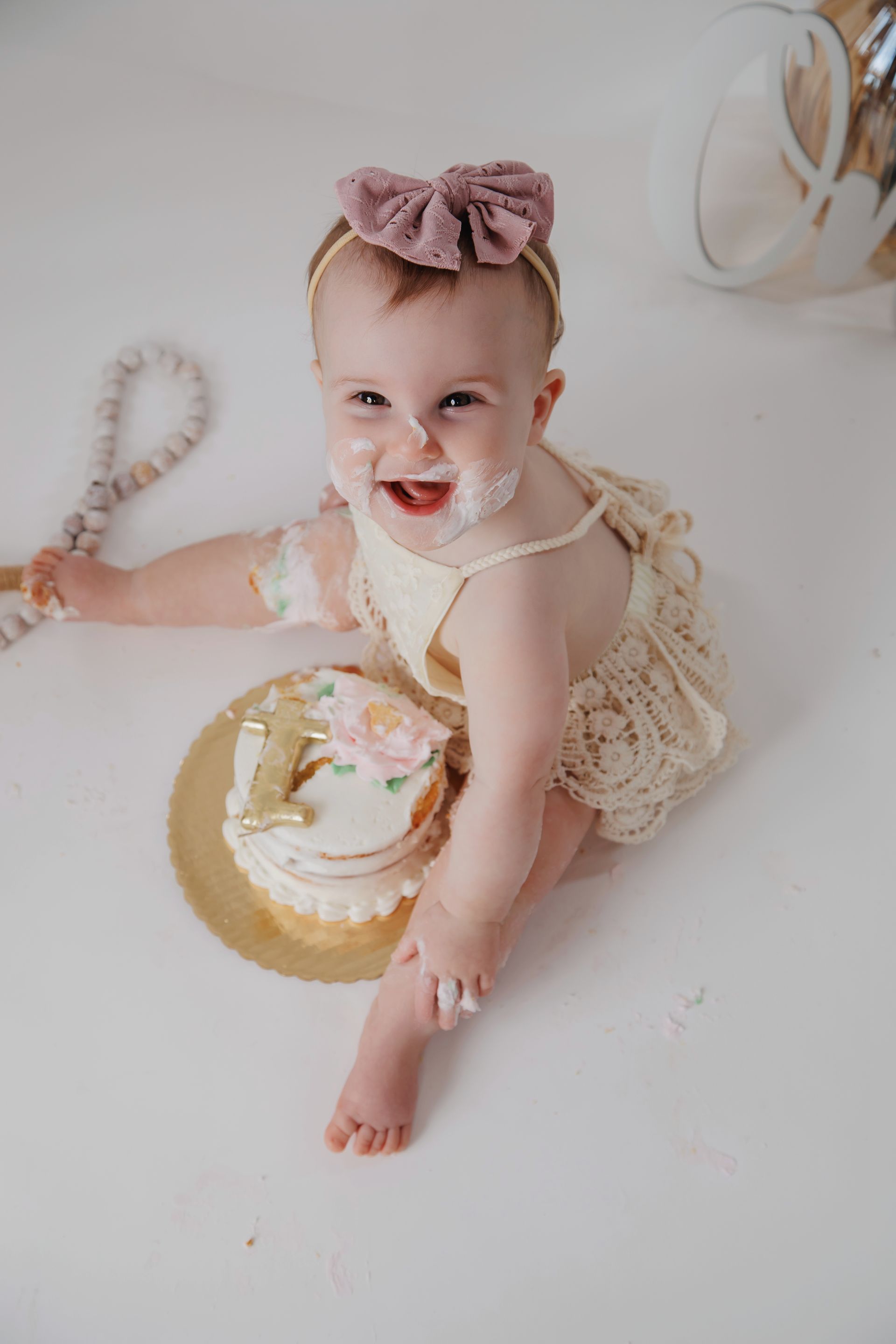 Smiling baby in a cream outfit with a mauve bow, sitting on a white blanket beside a gold cake.