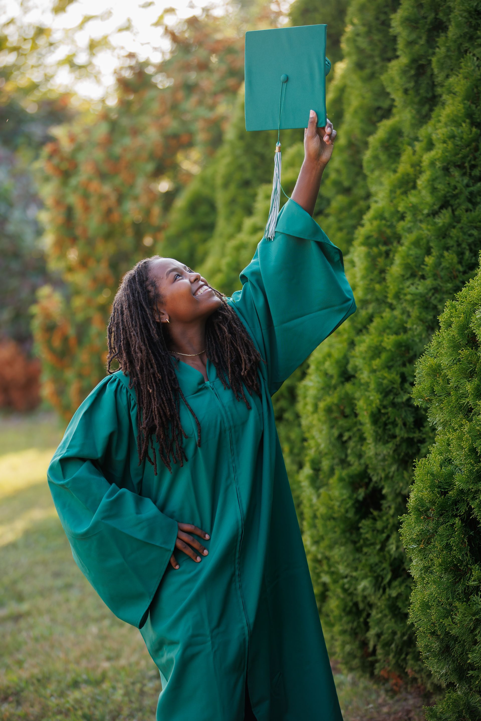 Person in a teal graduation gown reaching up to hold a matching cap outdoors by green trees