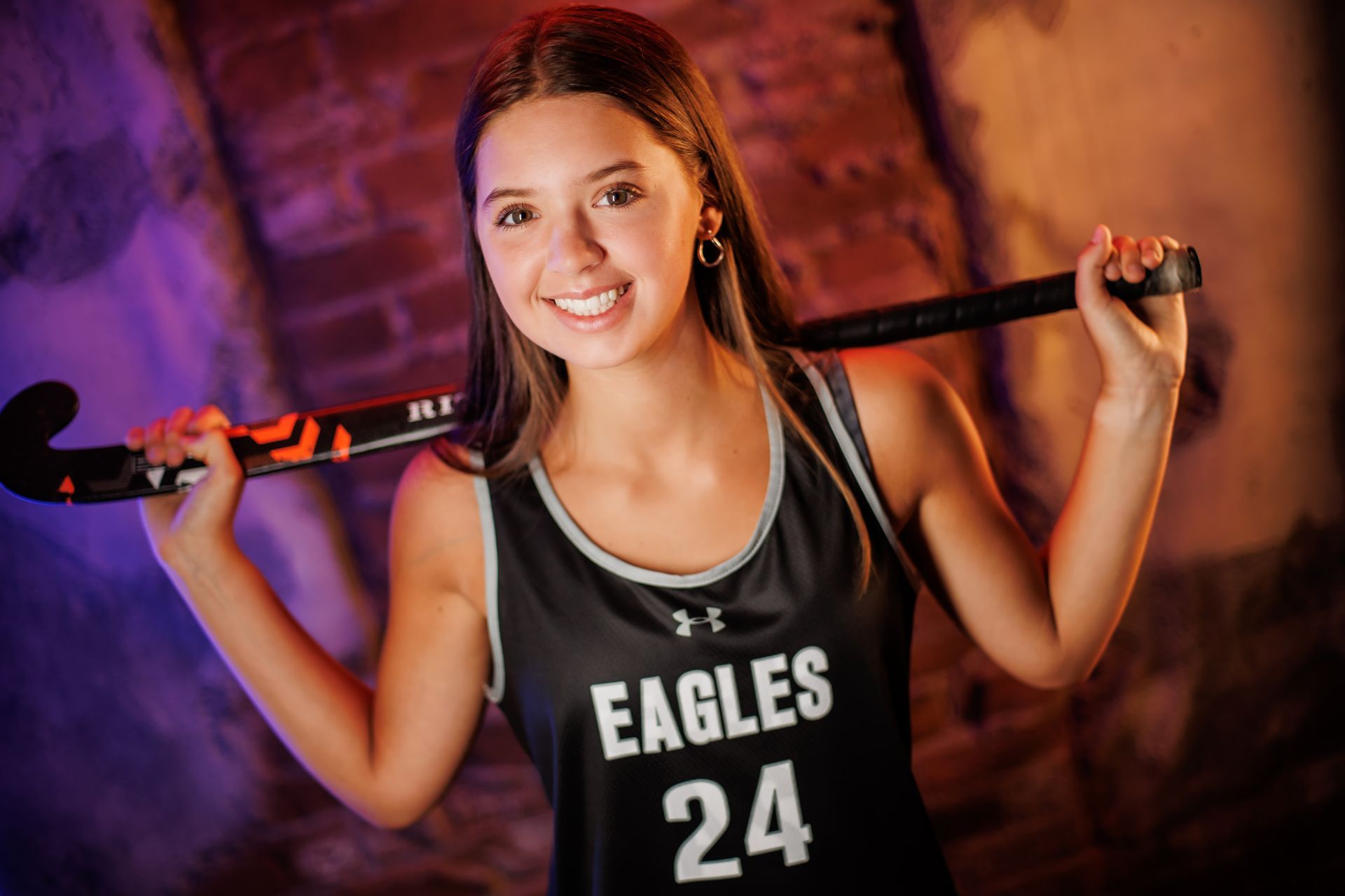 Smiling athlete in black EAGLES jersey holding a bat over her shoulders against a purple-lit background