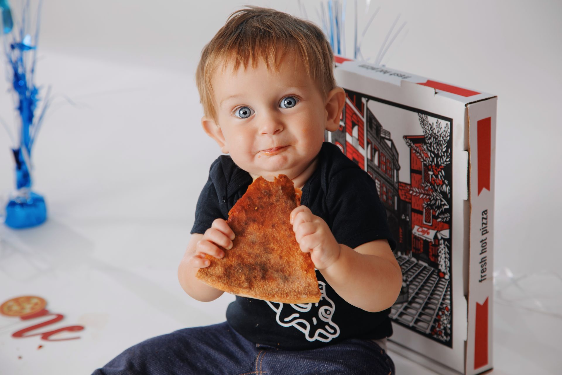 Toddler holding a slice of pizza beside a pizza box, sitting on a white surface.