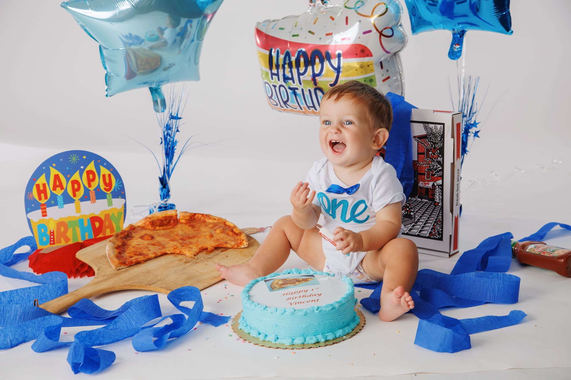 Baby smiling at a first birthday party with blue balloons, cake, pizza, and blue streamers