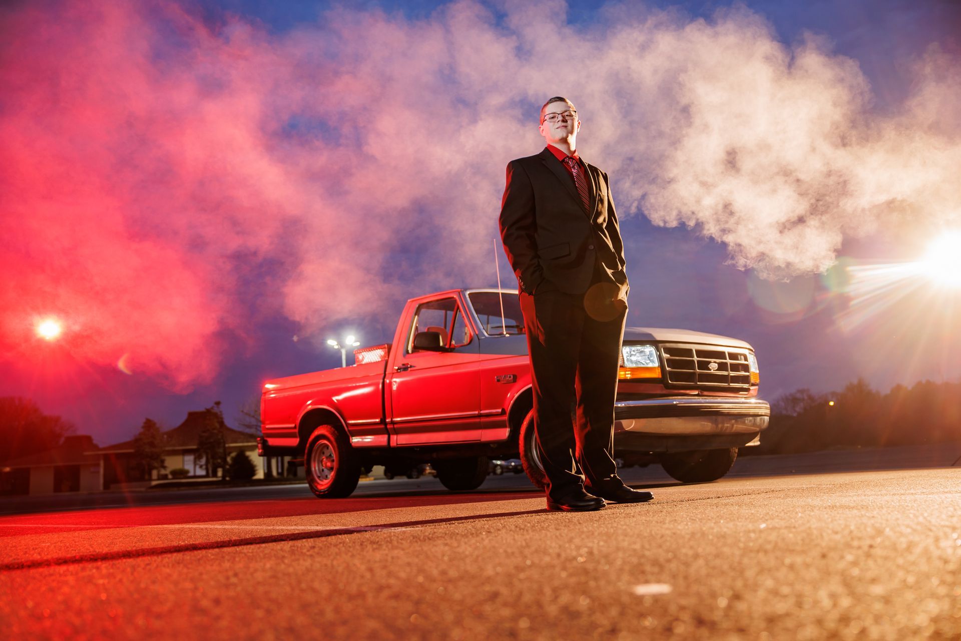 Man in suit standing by a red pickup truck at night with red smoke and bright headlights