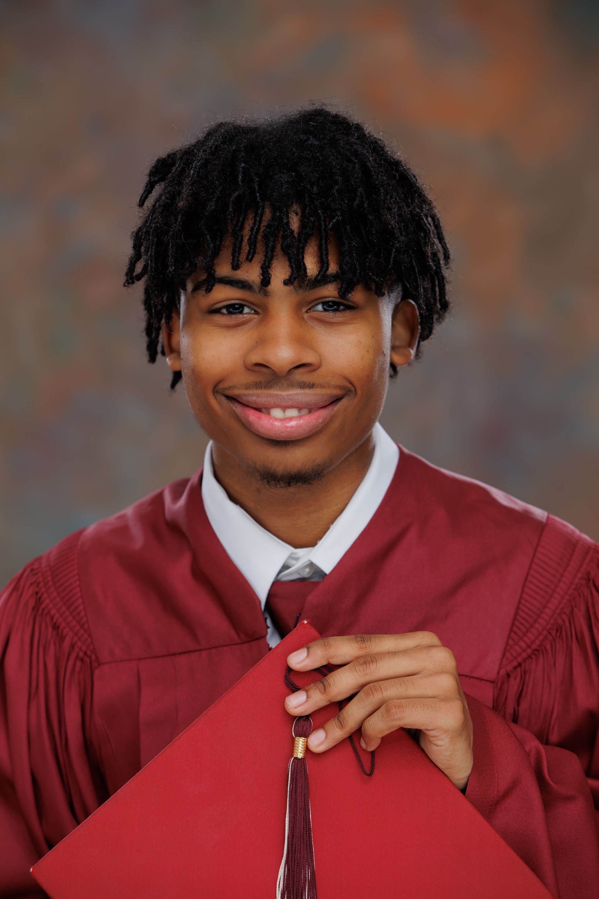 Graduate in red cap and gown smiling and holding a red diploma cover against a studio backdrop