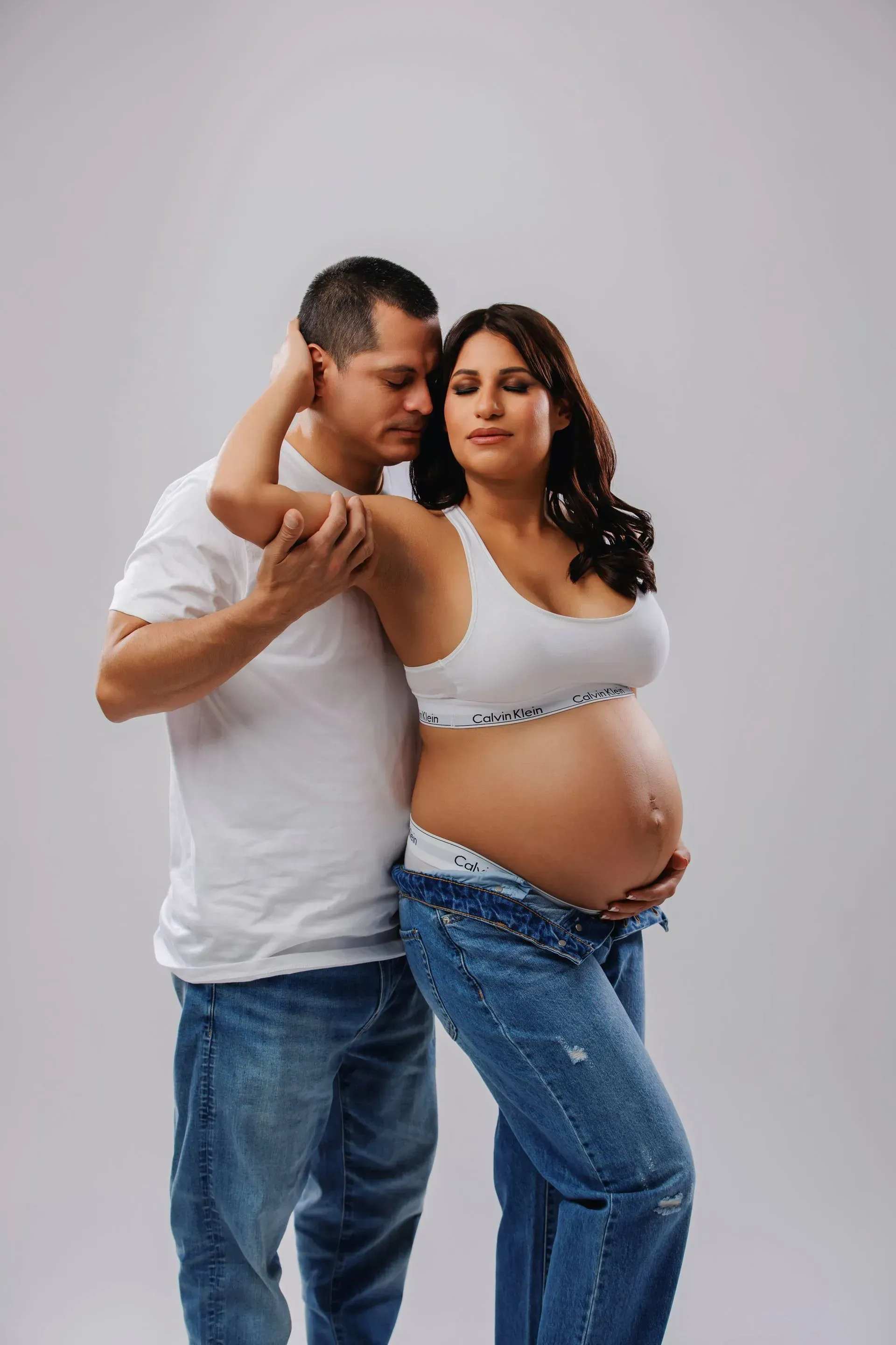 Pregnant couple posing against a light gray backdrop, standing close with arms around each other.