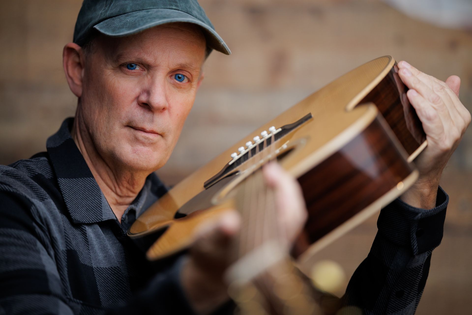 Man holding an acoustic guitar, wearing a cap and striped shirt, in a softly blurred indoor setting