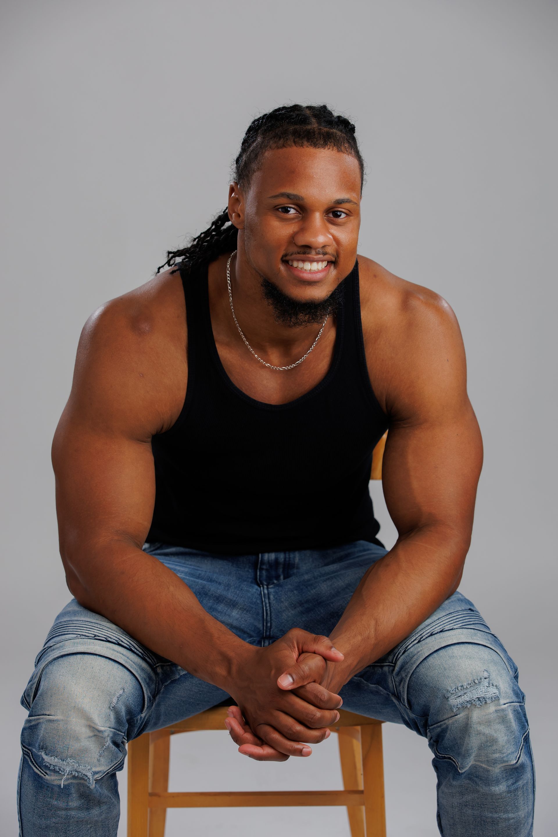 Smiling person in a black tank top and jeans sitting on a wooden stool against a gray background