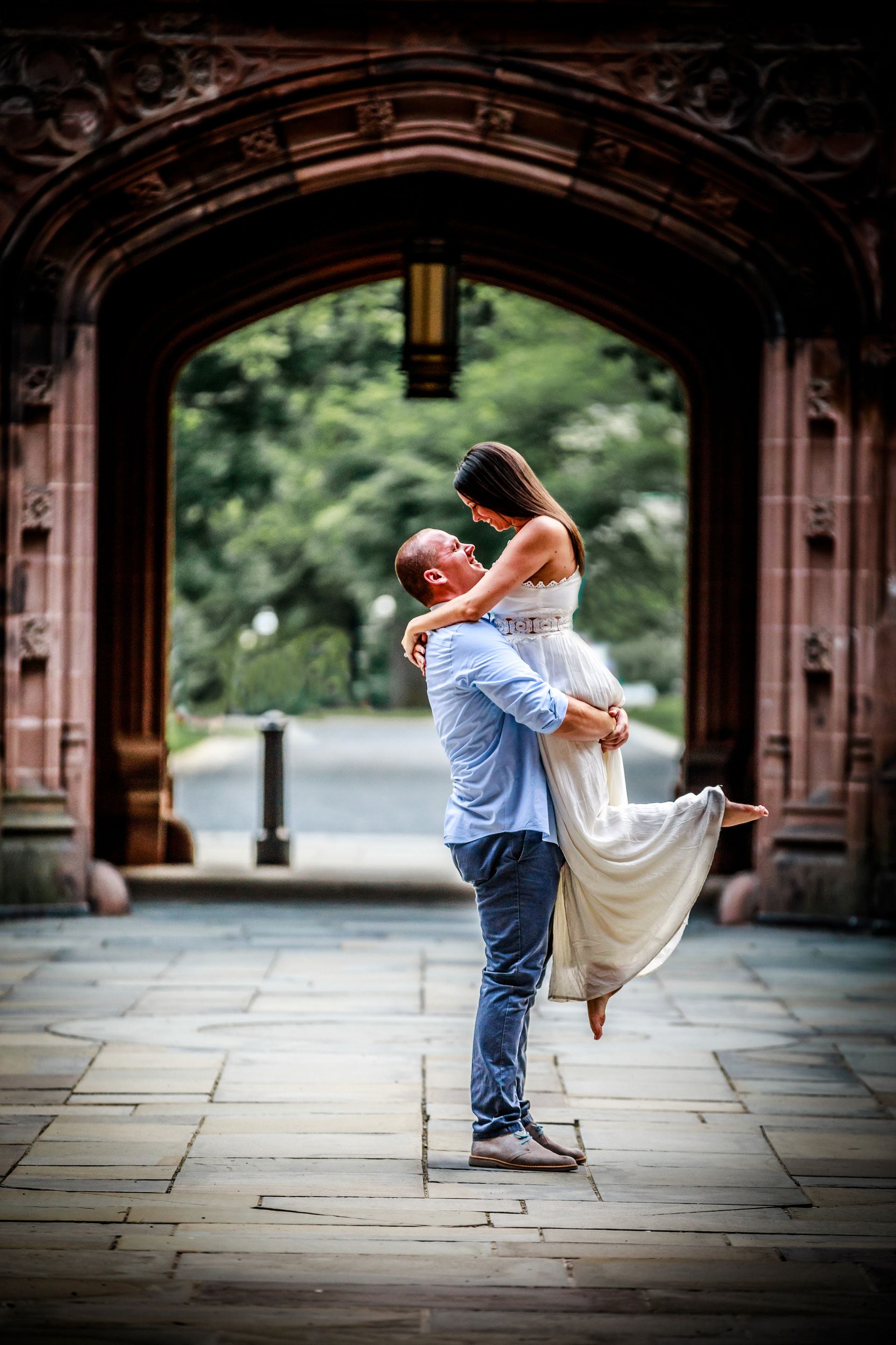 Couple embracing and lifting each other in a stone archway, with greenery blurred in the background.