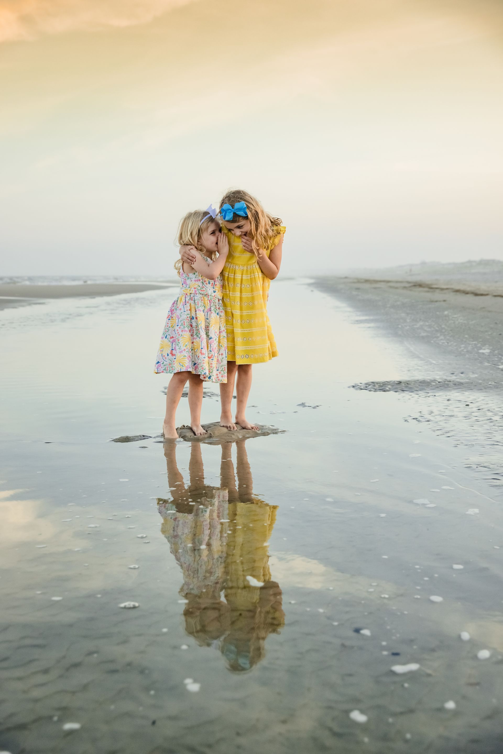 Two children in colorful dresses standing close together on a wet beach at sunset