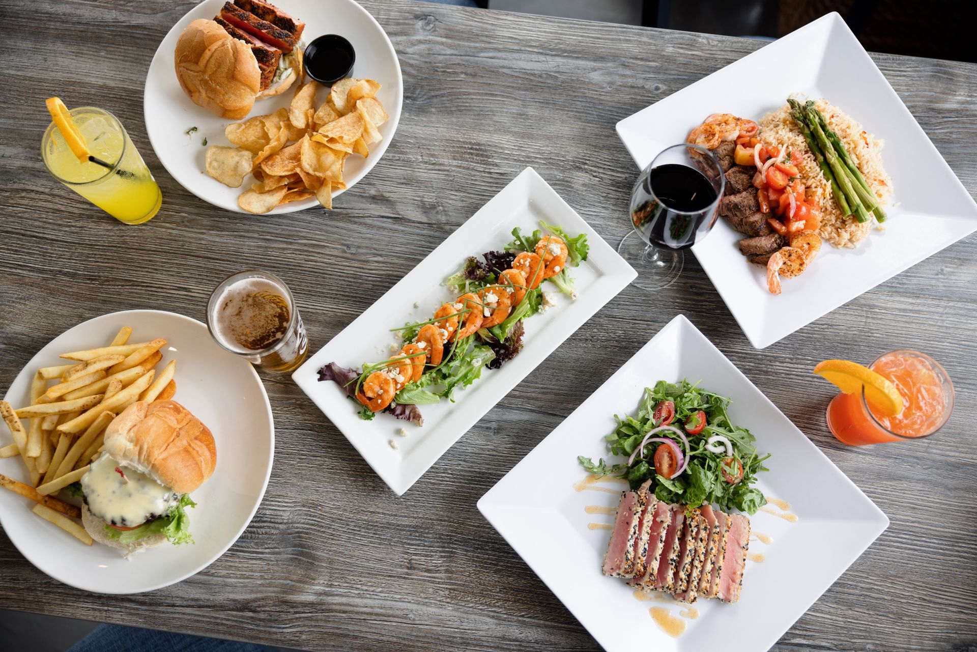 Assorted sushi and fried dishes arranged on white plates on a gray table, with drinks and sauces