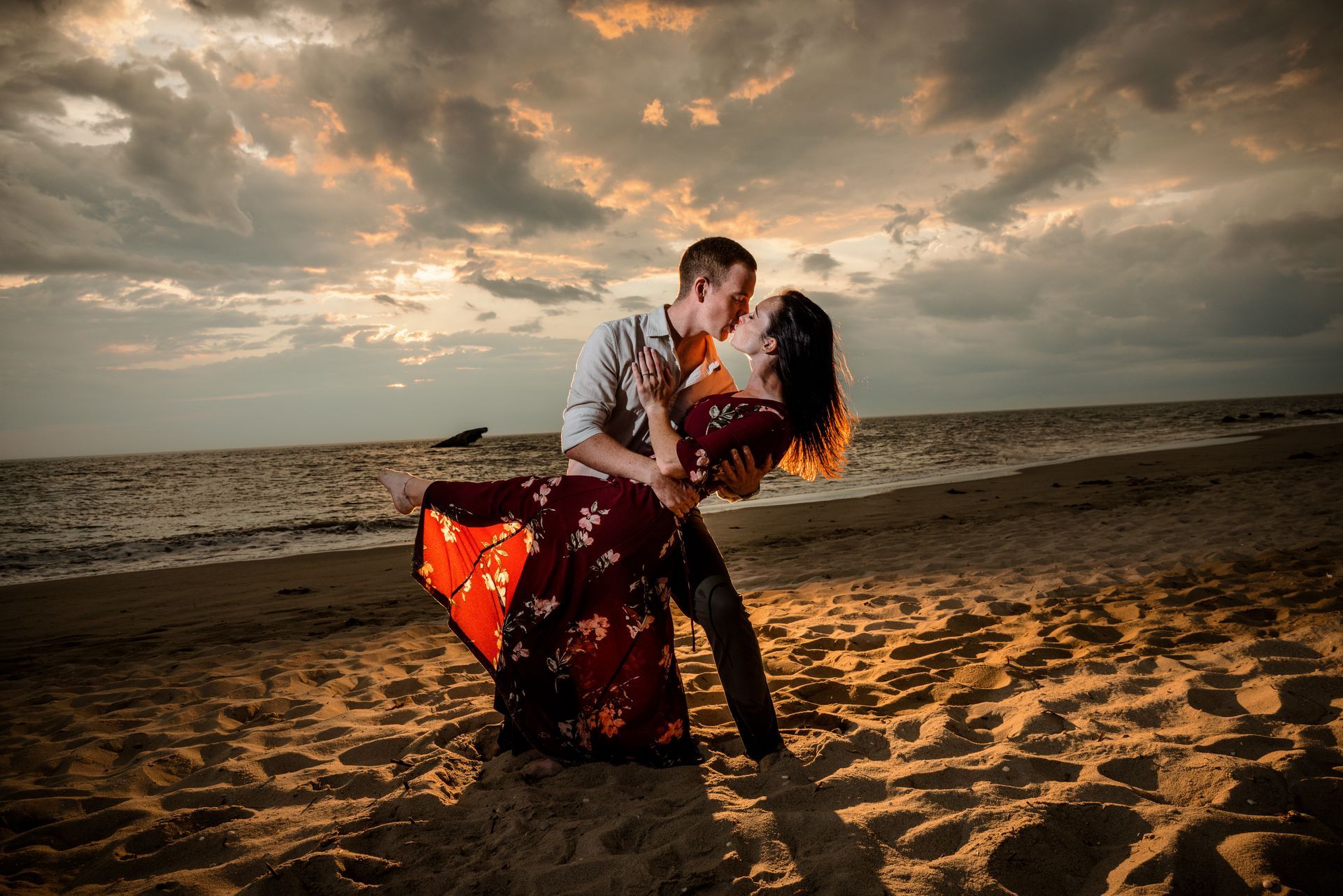 Couple embracing on a beach at sunset, with dramatic clouds and a red flowing dress