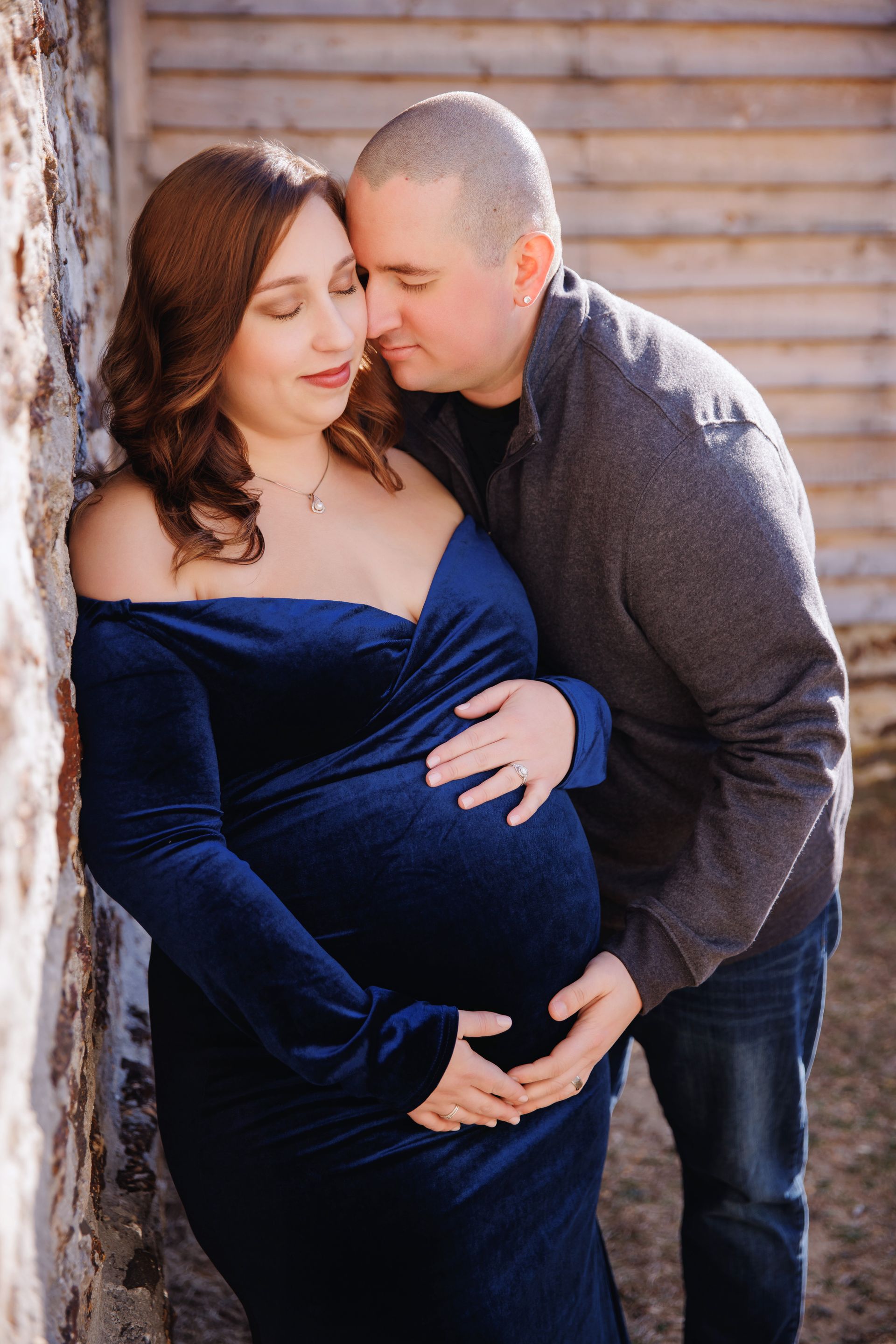 Couple posing outdoors, embracing with hands on a pregnant belly, in front of a wooden fence