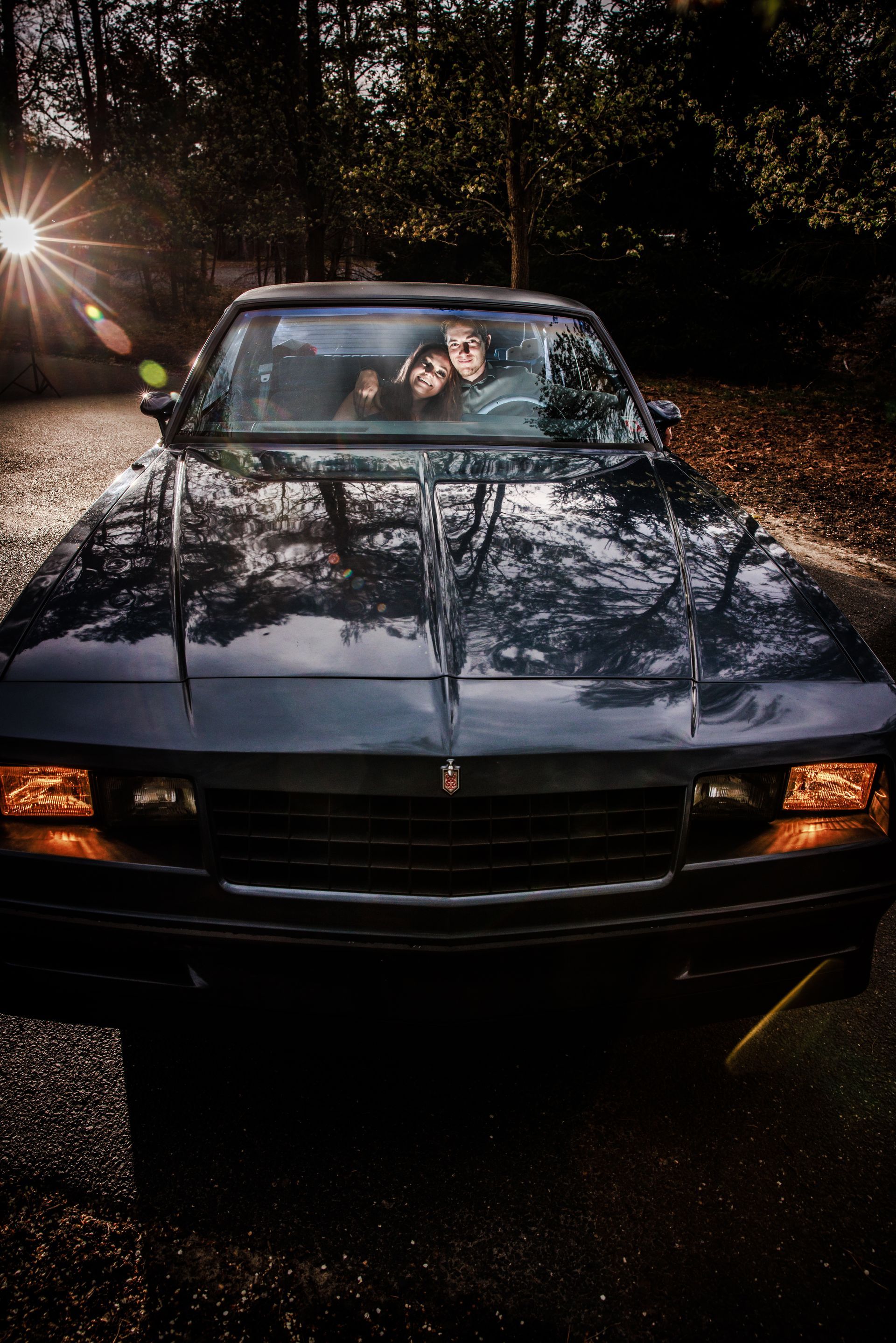 Two people in a dark blue pickup truck on a tree-lined road at night, with headlights and reflections on the hood.