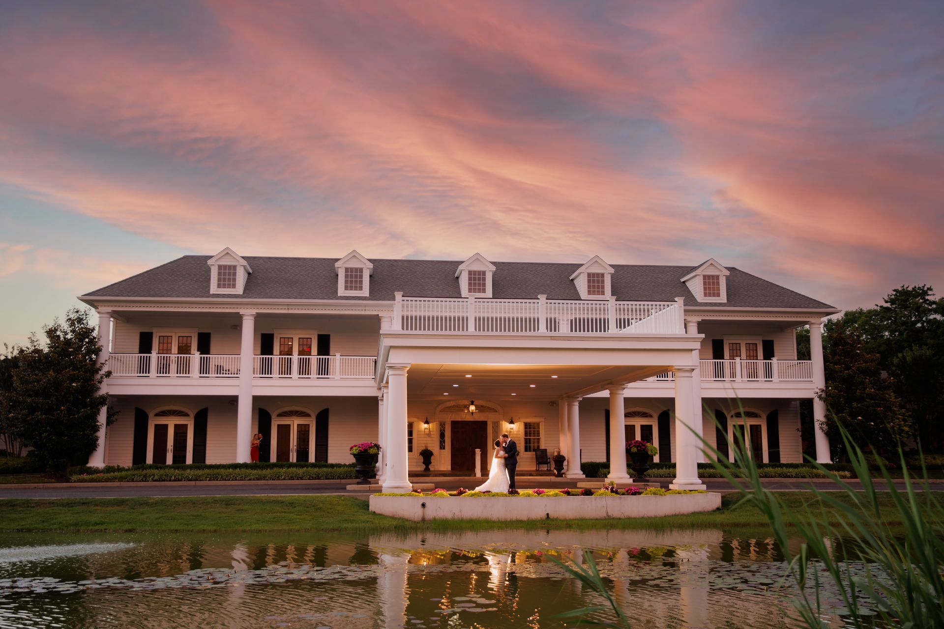 Large white mansion with a lit portico reflected in a pond at sunset