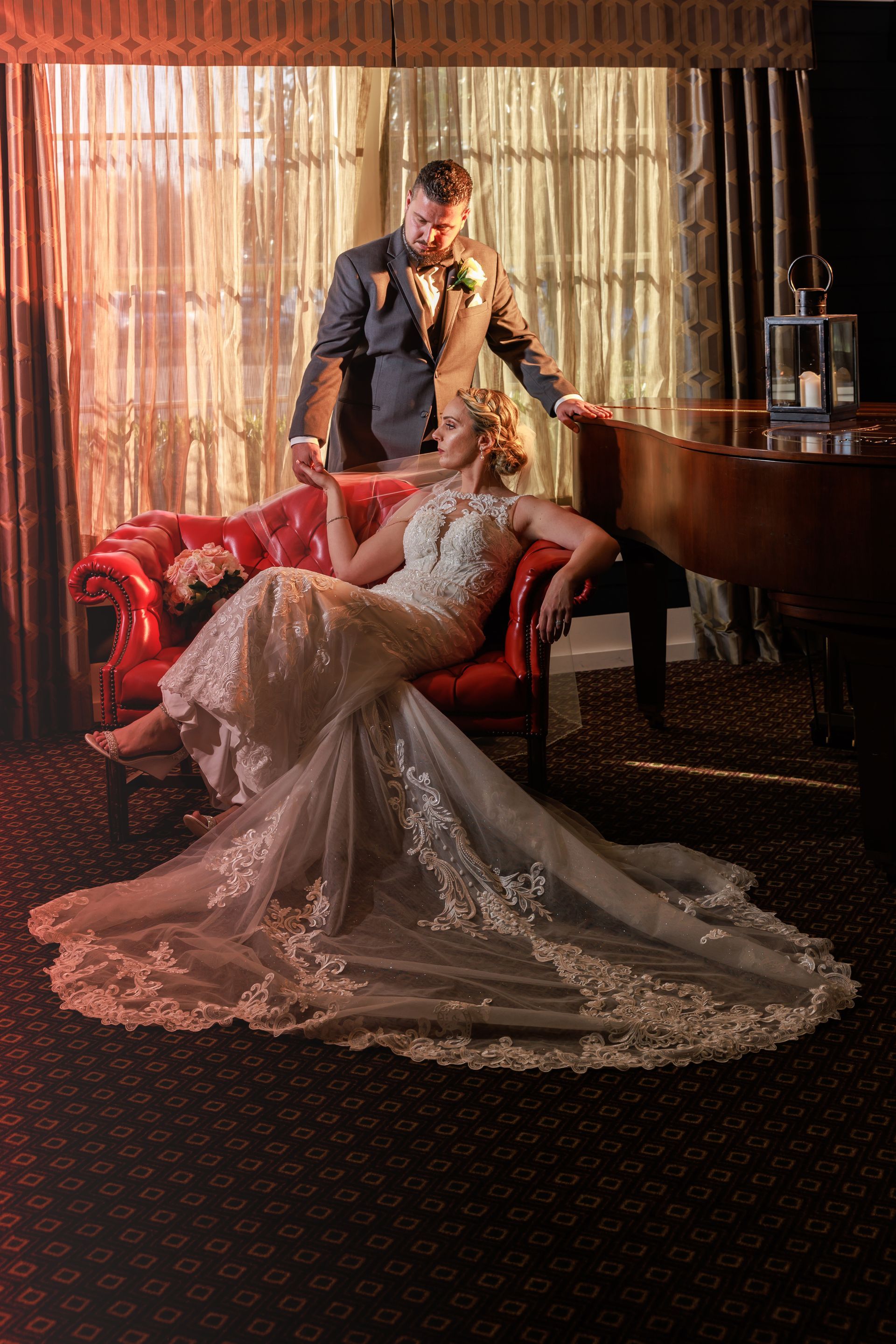 Bride on a red sofa with groom standing beside her in a warmly lit room, wedding dress trailing across the floor