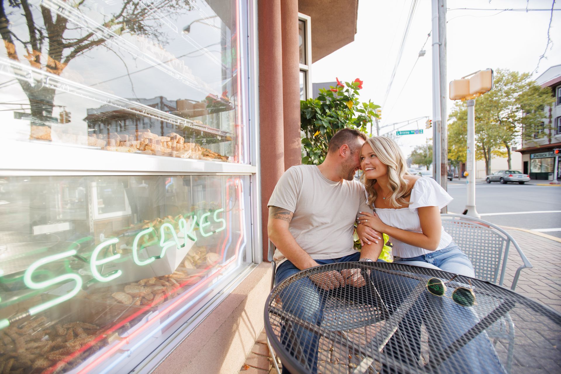 Couple leaning close at a café table by a window, with street view and “Sea Cakes” sign reflected beside them