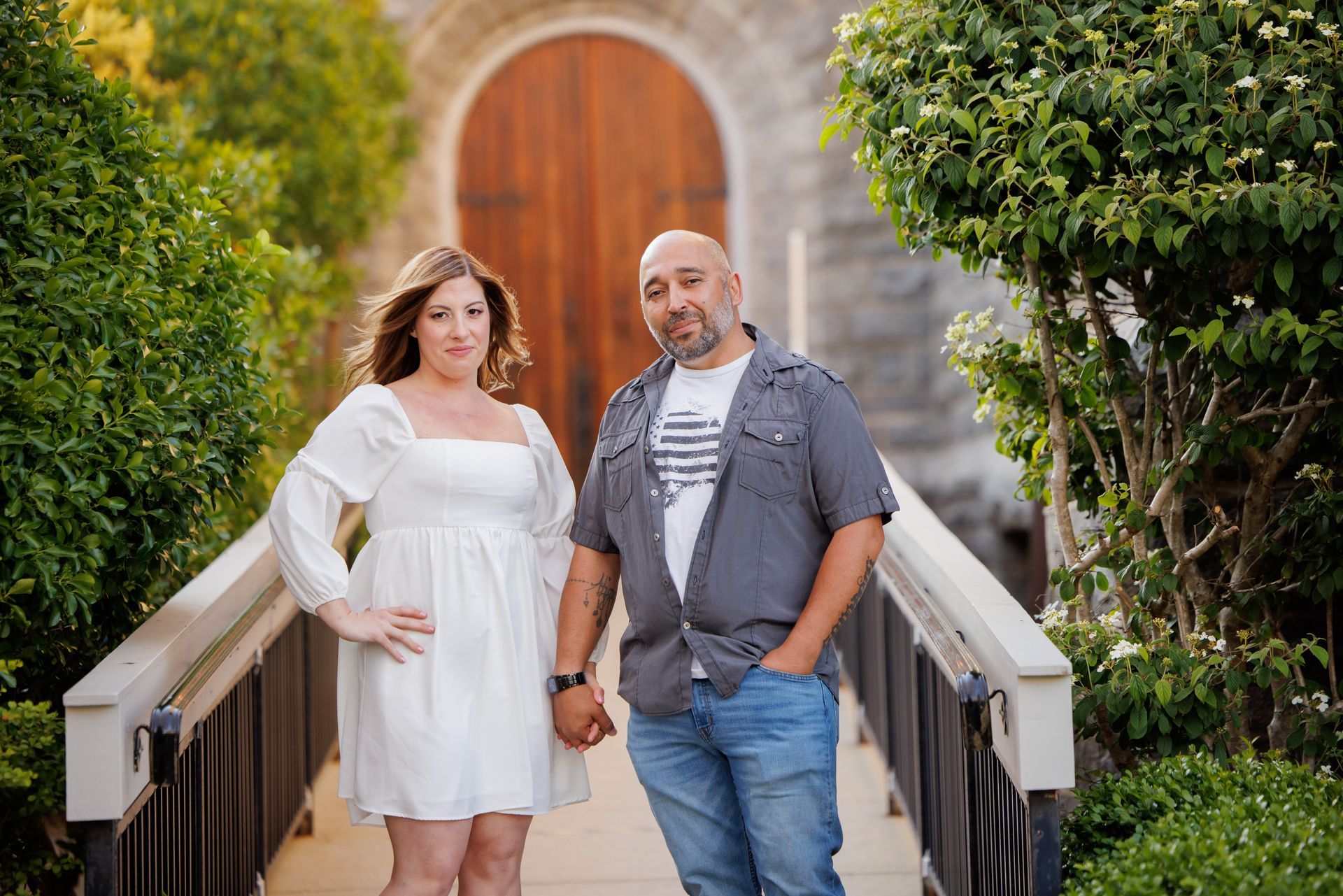 Two people standing on a wooden bridge in front of an arched stone doorway and greenery