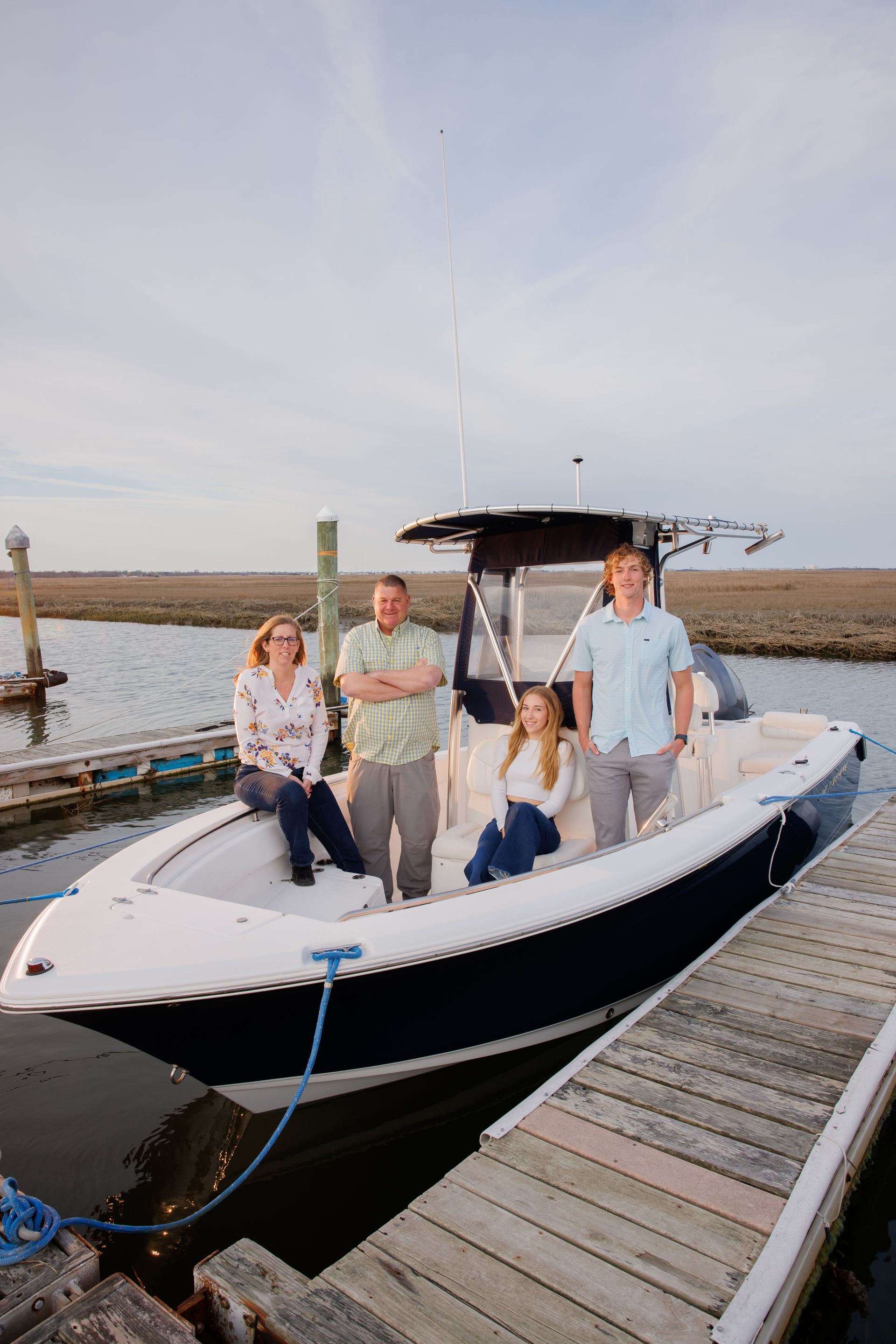 Four people standing on a white motorboat docked at a marina under a cloudy sky