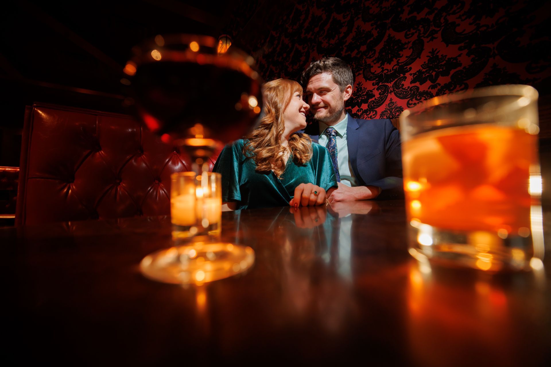 Couple sharing a candlelit drink in a dim bar, with warm amber glasses in the foreground