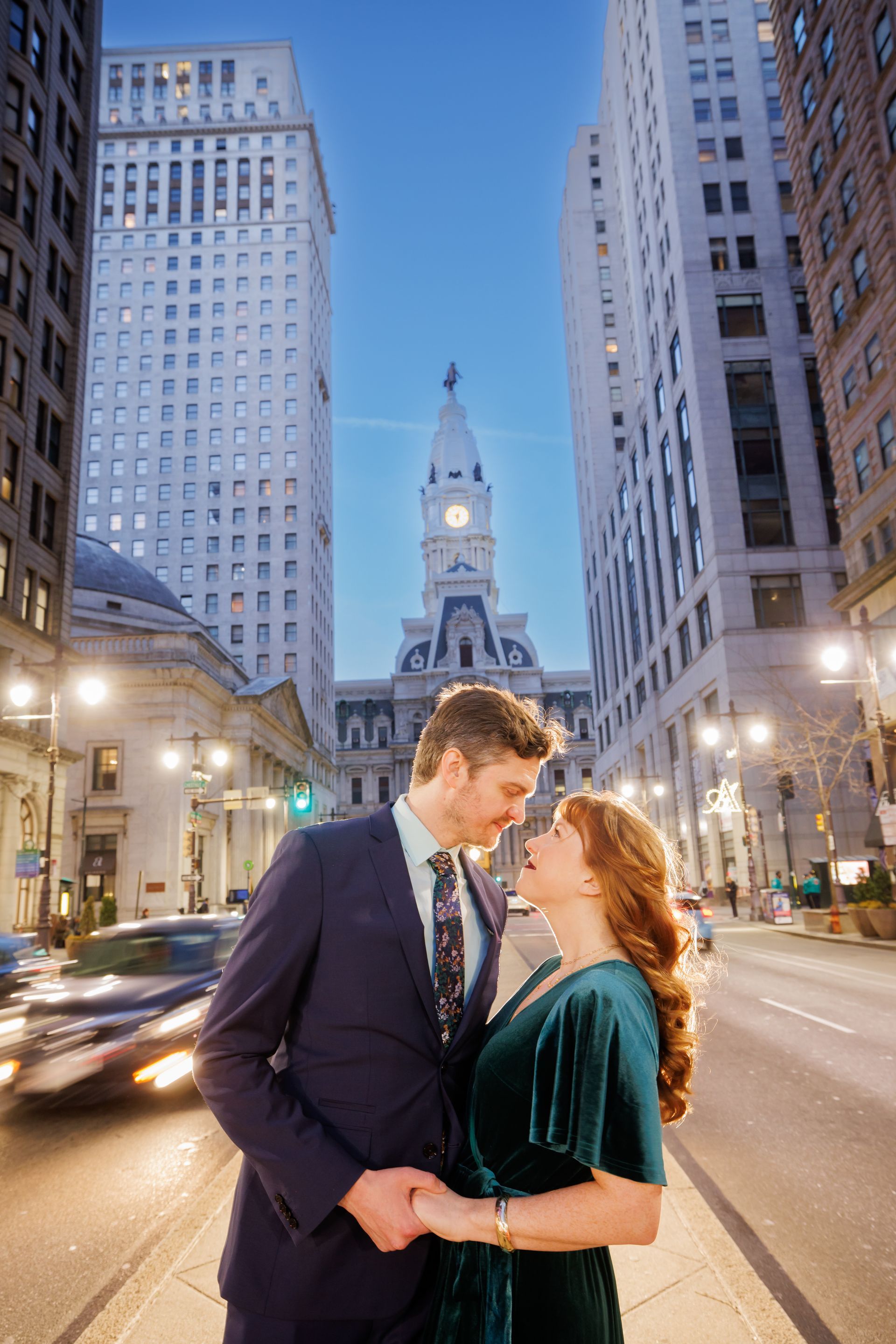 Couple embracing on a city street at dusk, framed by tall buildings and a lit clock tower behind them.
