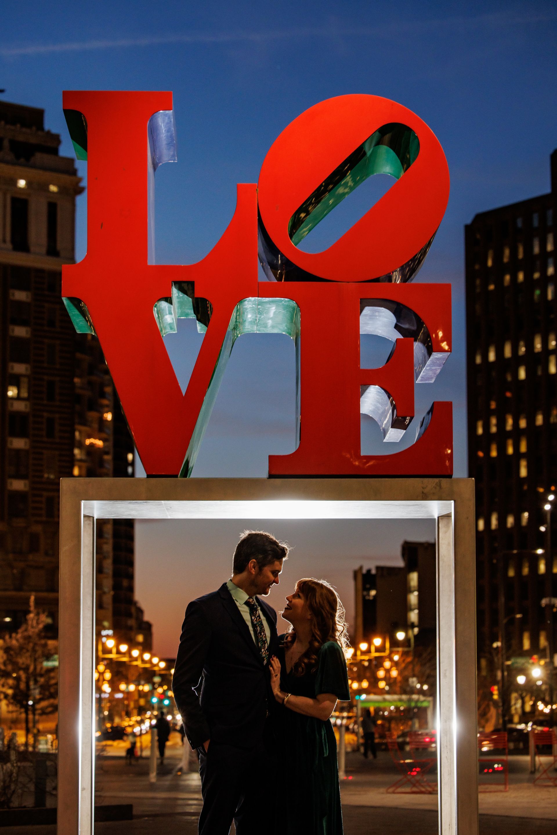 Red LOVE sculpture above a couple standing in a lit frame at night in a city square