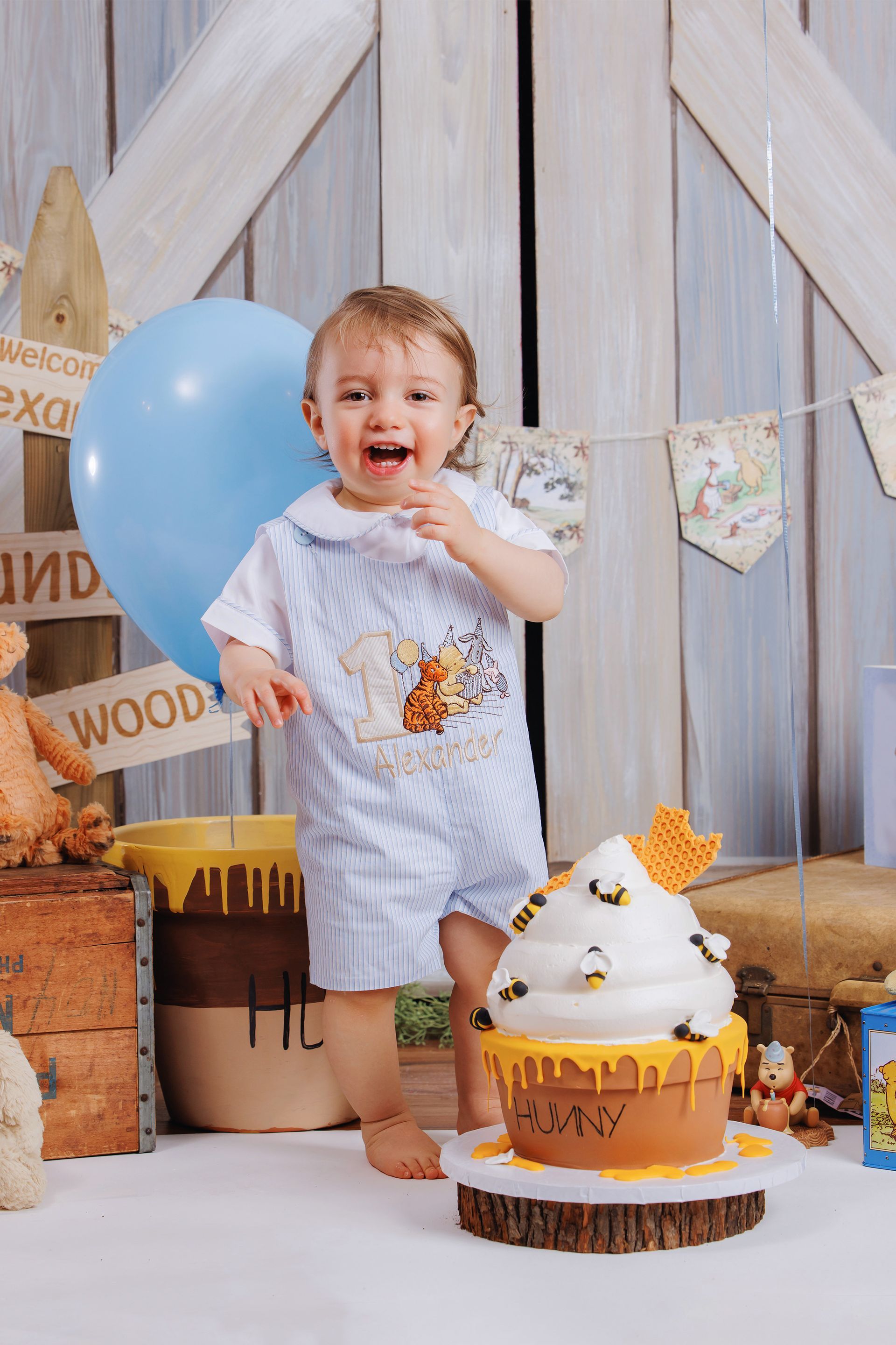 Toddler smiling beside a white-and-gold birthday cake, with a blue balloon and rustic party decor