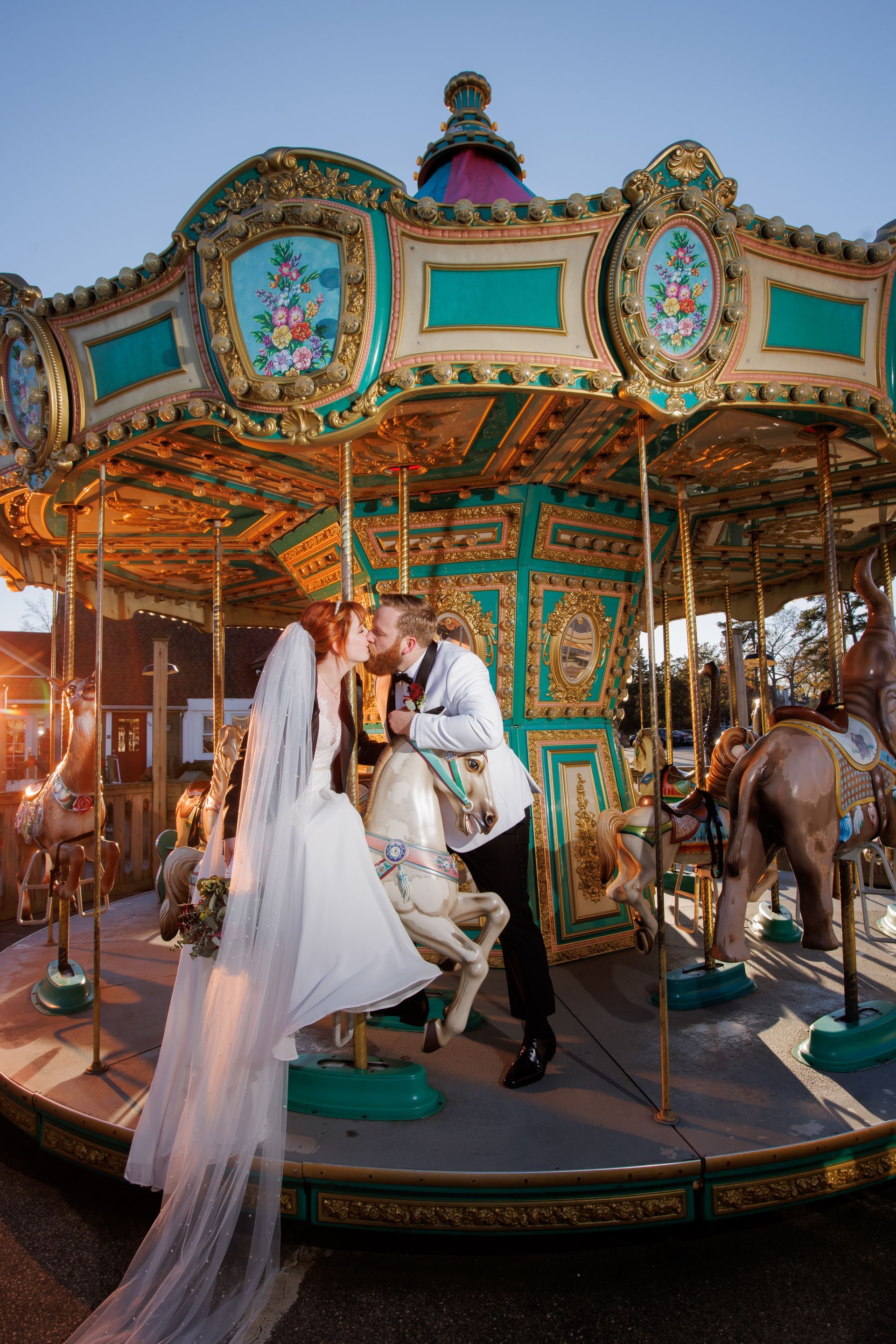 Couple in wedding attire on a decorated carousel at sunset, with white horse figures and ornate gold trim