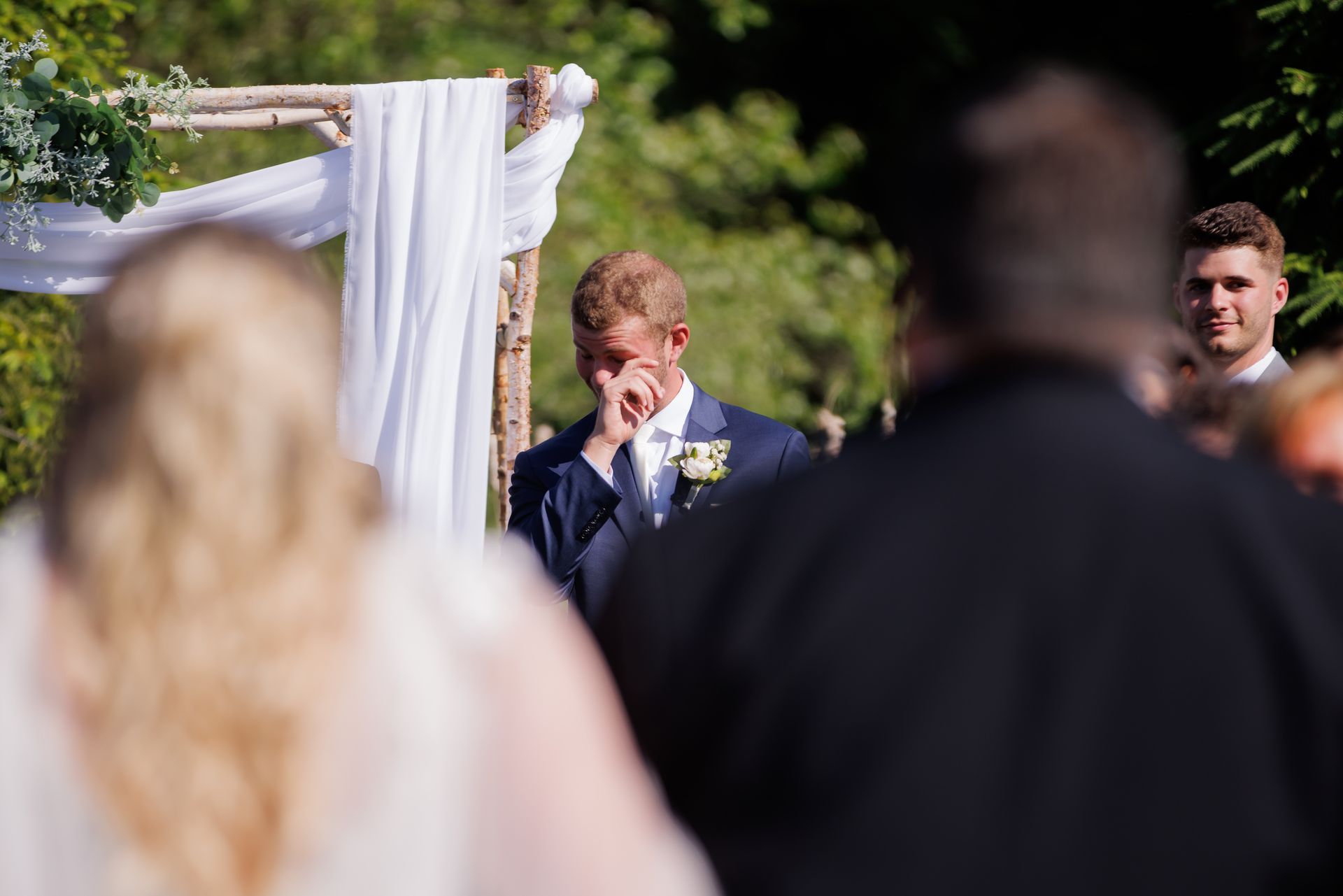 Outdoor wedding ceremony with groom wiping his eye as guests watch under a white floral arch