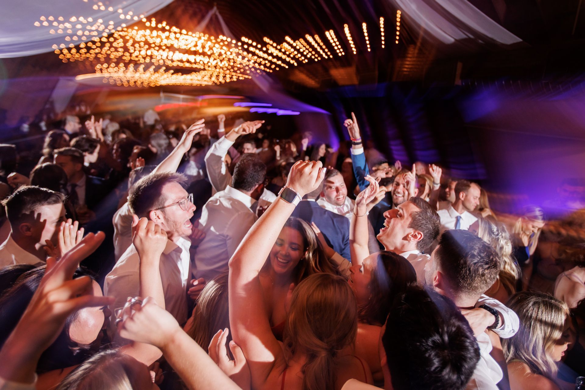 Crowded nightclub dance floor with people dancing under purple and warm ceiling lights