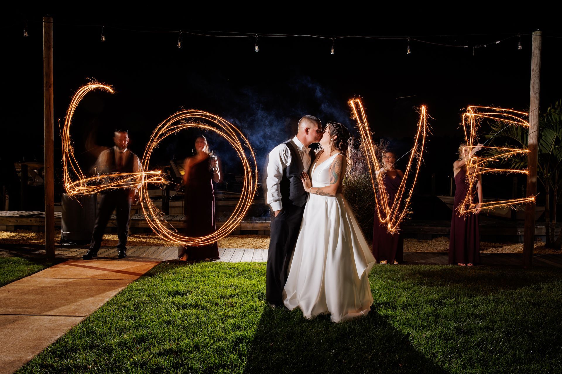 Couple in wedding attire standing under sparkler-lit “LOVE” letters at night outdoors