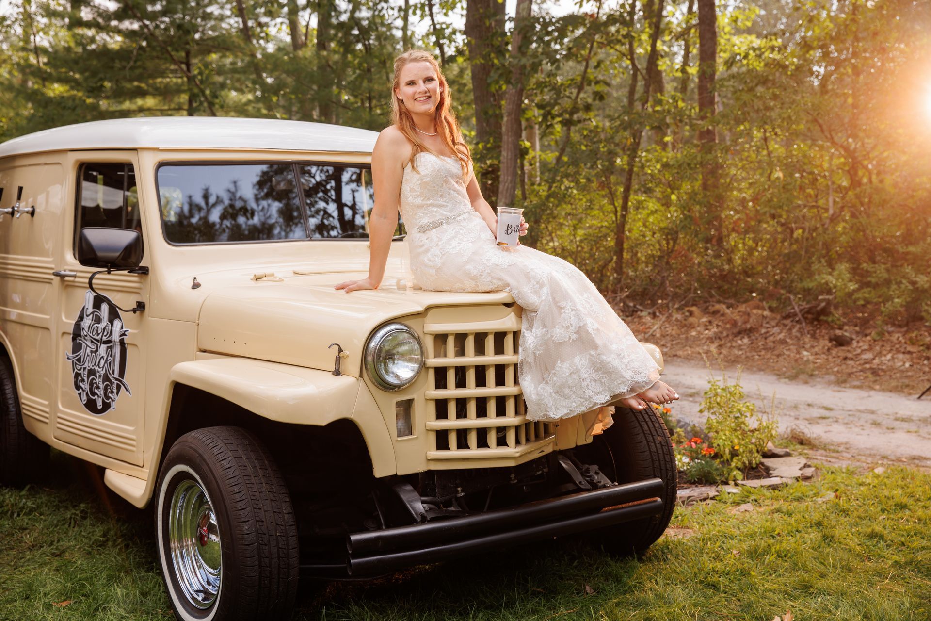 Bride in a white dress sitting on a beige vintage truck in a sunny wooded setting