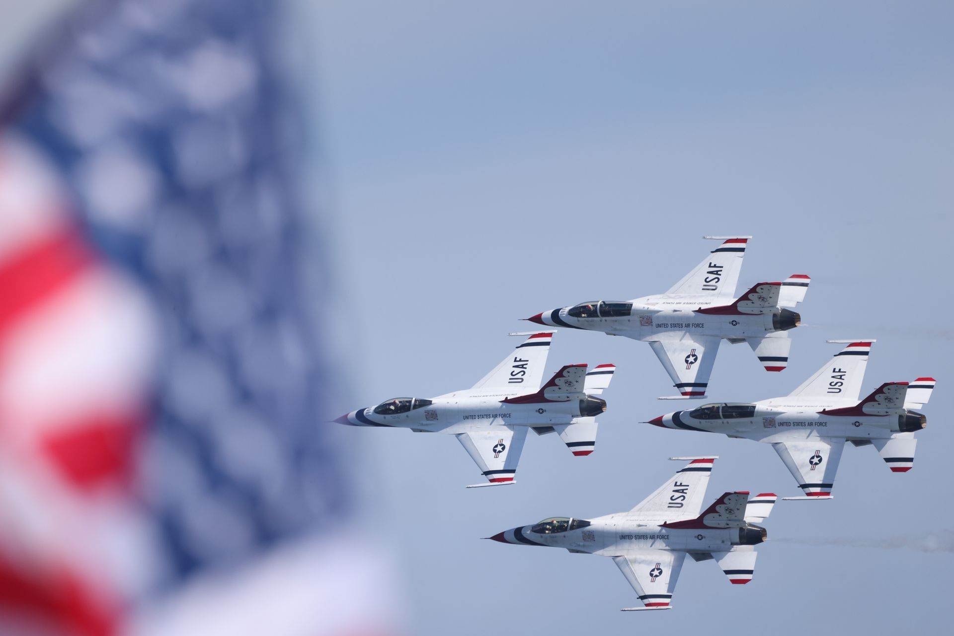 Four fighter jets fly in formation against a blue sky, with a blurred American flag in the foreground.