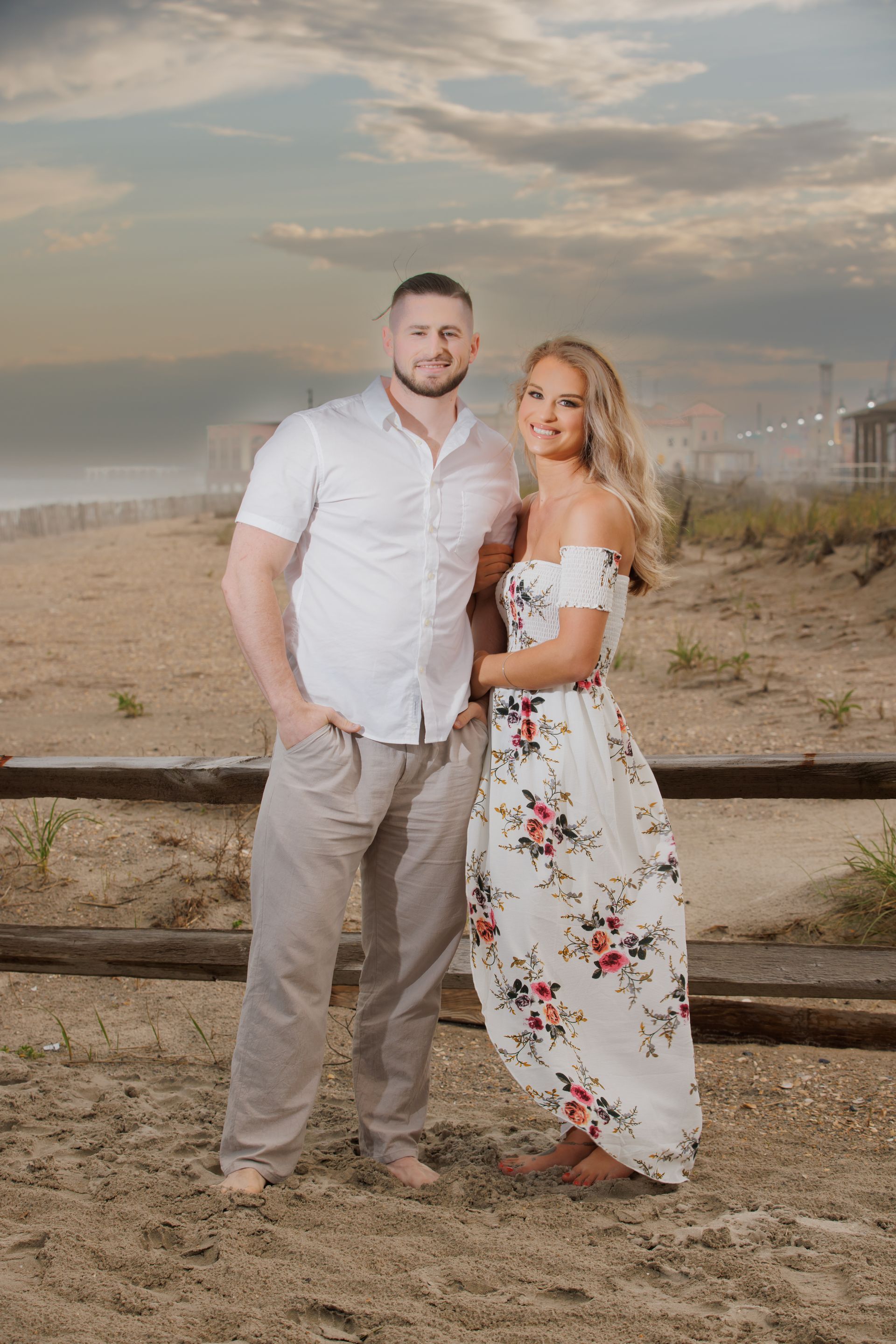 Couple standing on a sandy beach at sunset, with a wooden fence and cloudy sky behind them