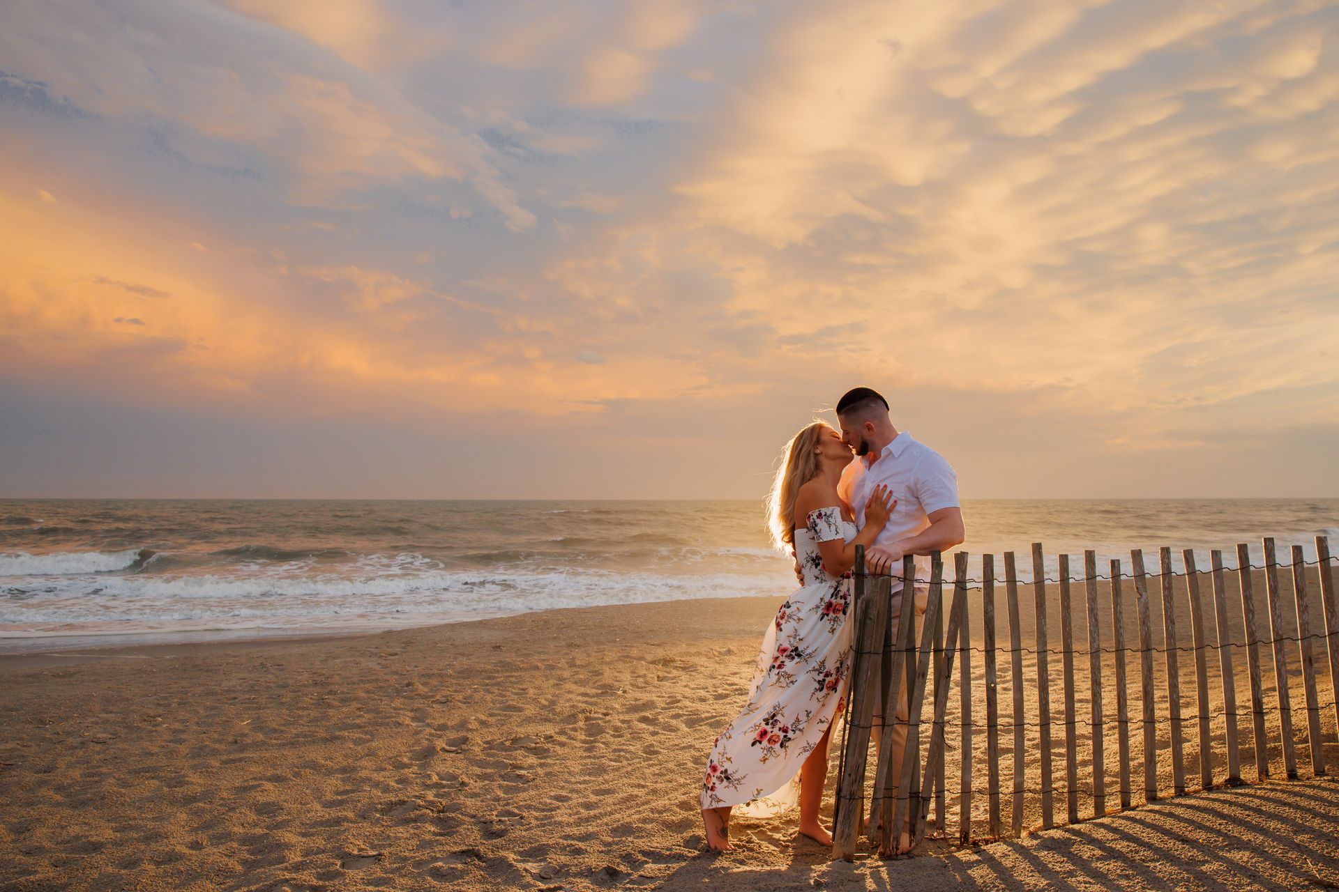 Couple embracing by a fence on a beach at sunset, with warm pastel sky and waves in the background