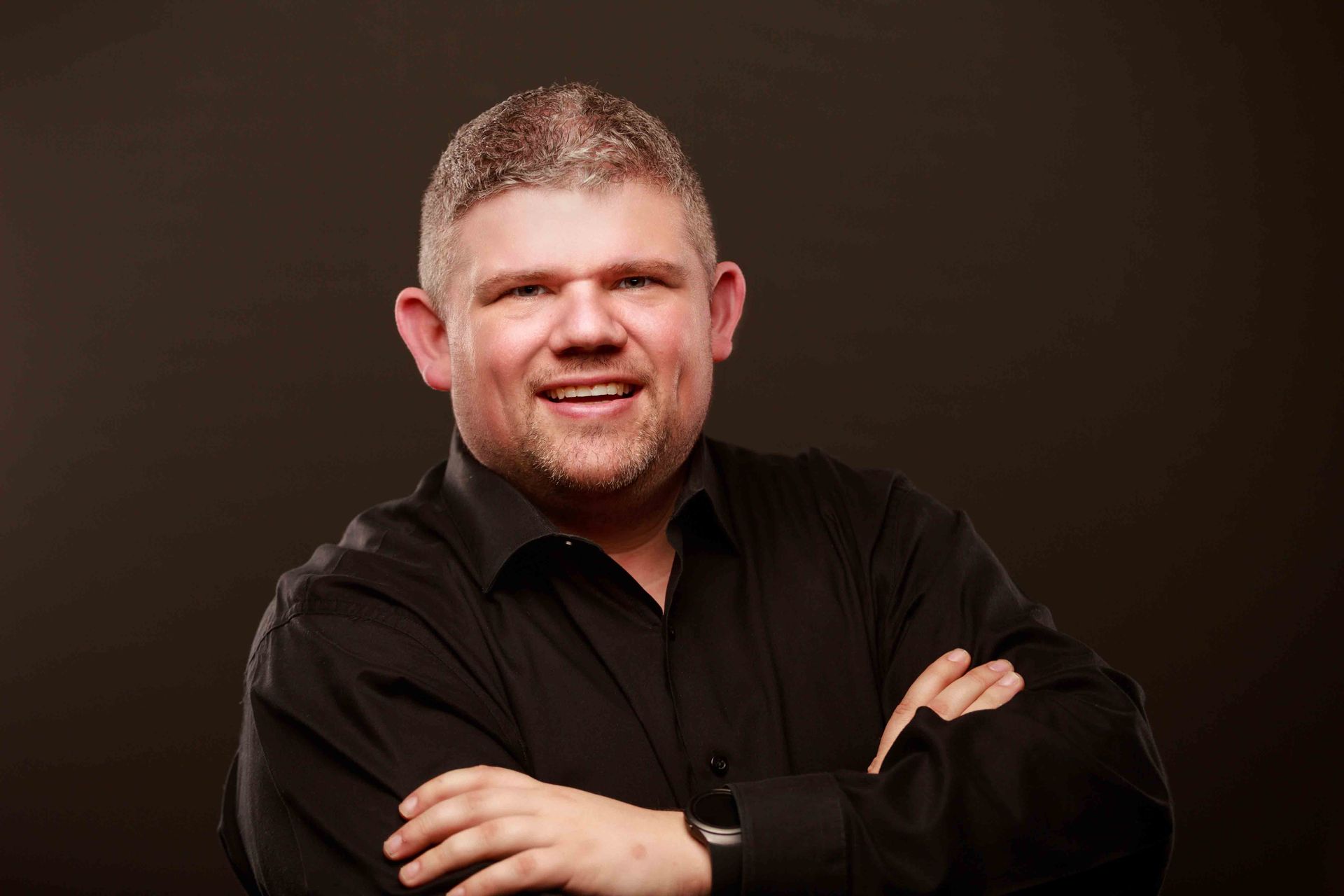 Smiling person in a black shirt with arms crossed against a dark brown studio background