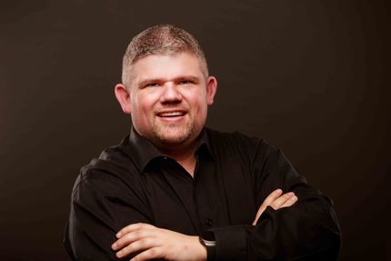 Smiling person in a black shirt with arms crossed against a dark brown studio background