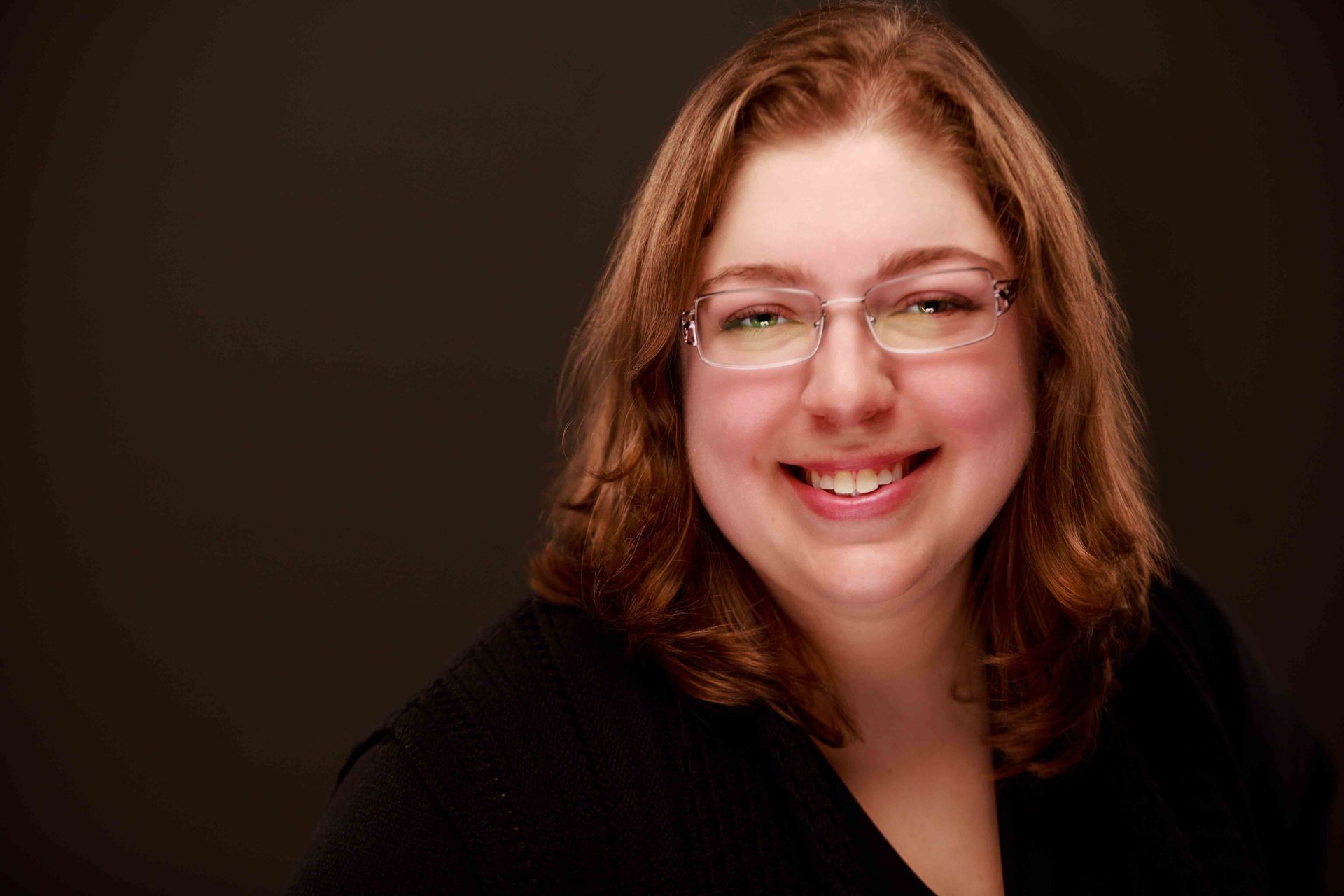 Smiling person with glasses and shoulder-length brown hair against a dark background