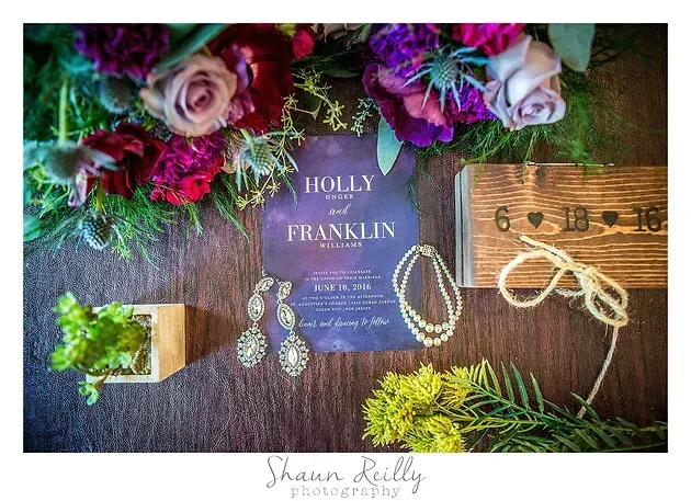 Wedding flat lay with purple invitation, flowers, jewelry, rope, and wooden box on a grass background