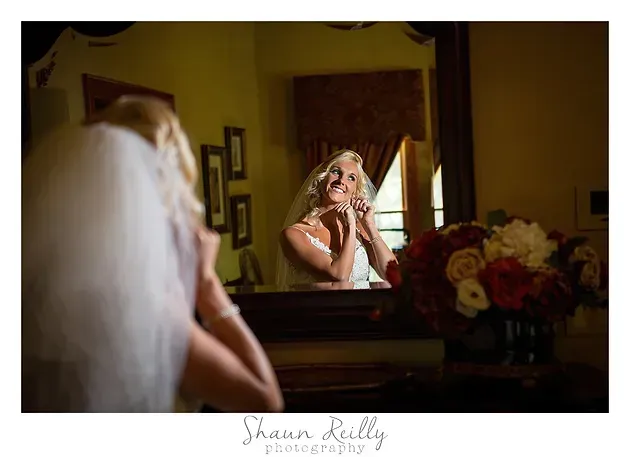 Bride adjusting veil while looking in a mirror, with soft light and flowers in the foreground