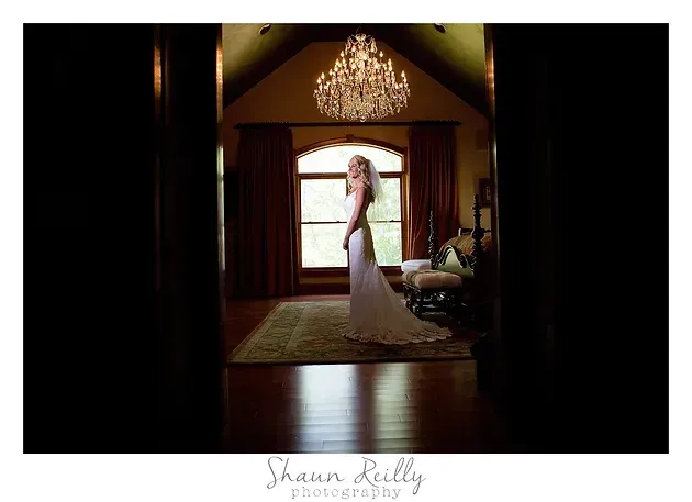 Bride in a white gown standing in a warmly lit doorway beneath a chandelier