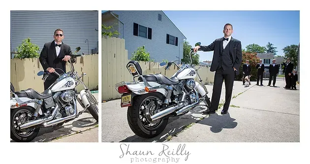 Man in black suit standing beside a silver motorcycle outdoors at an event, with another bike nearby